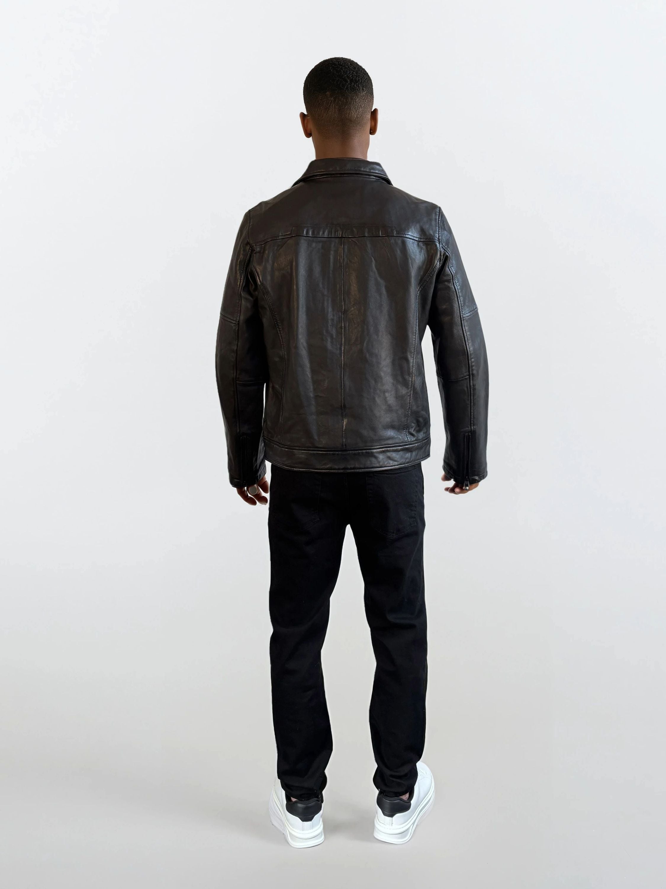 Eine Person trägt eine schwarze Herren Lederjacke aus Schafsleder mit Ärmel-Reißverschlüssen von trueprodigy, dazu schwarze Hosen und weiße Sneaker vor einem schlichten, hellen Hintergrund.