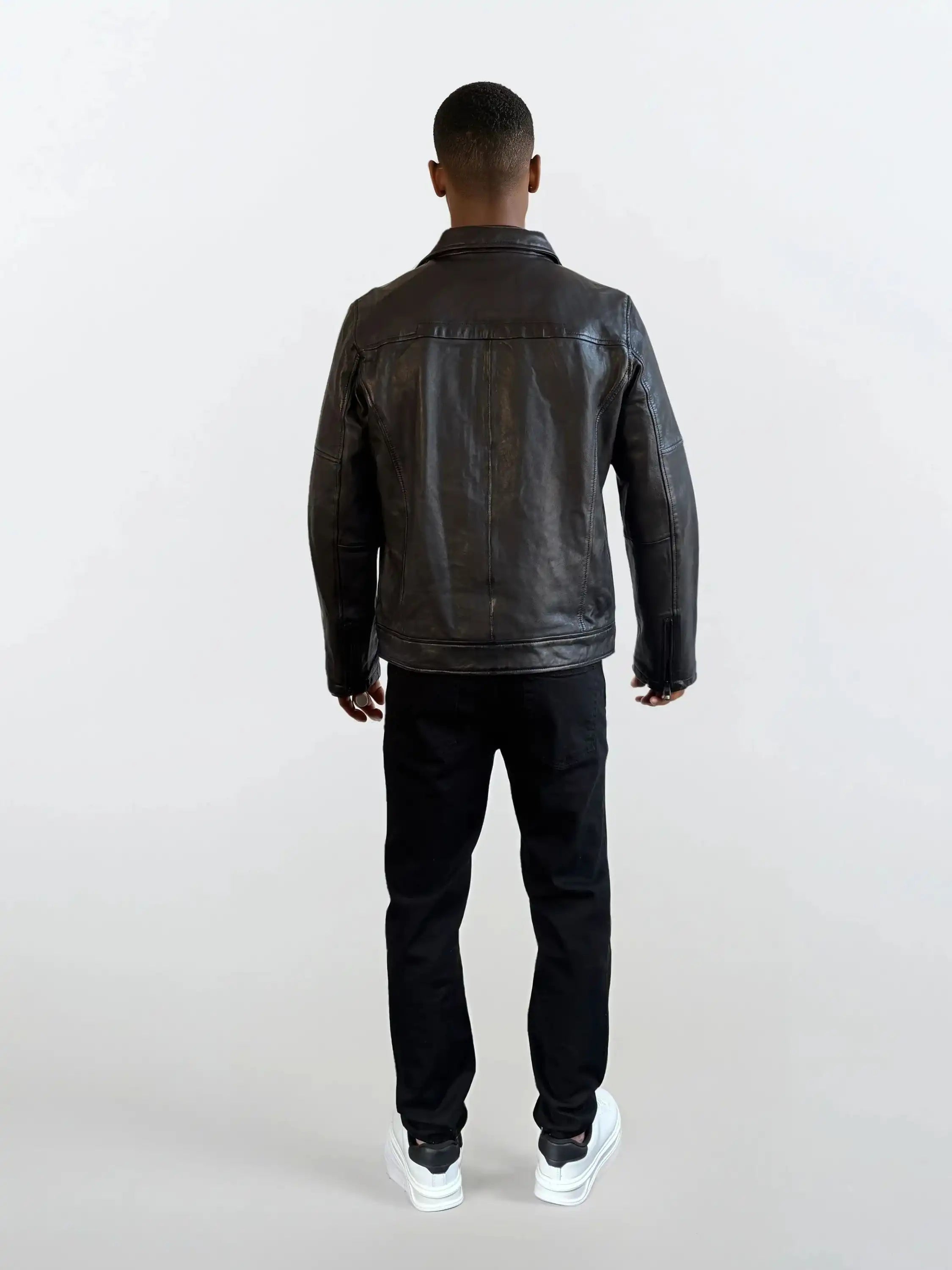 Eine Person trägt eine schwarze Herren Lederjacke aus Schafsleder mit Ärmel-Reißverschlüssen von trueprodigy, dazu schwarze Hosen und weiße Sneaker vor einem schlichten, hellen Hintergrund.