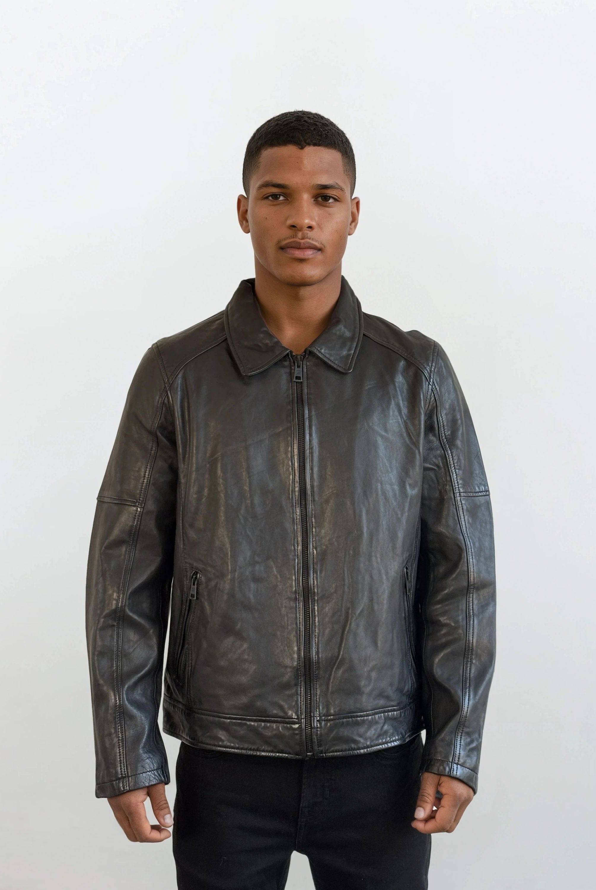 Vor weißem Hintergrund trägt eine Person die trueprodigy Herren Lederjacke aus Schafsleder in Schwarz mit Ärmel-Reißverschlüssen über schwarzem Shirt und Hose und blickt neutral in die Kamera.