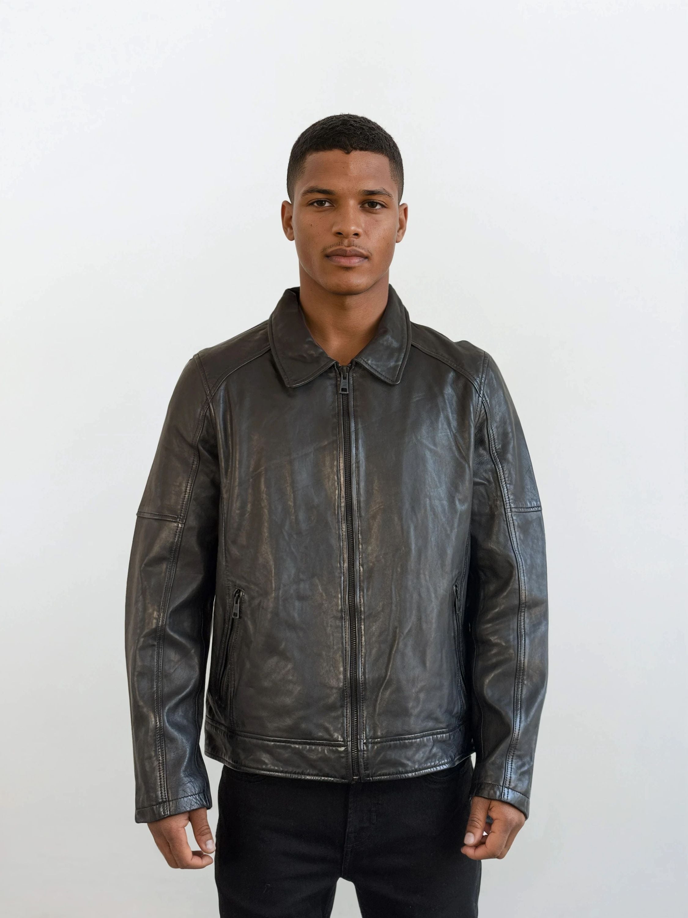Vor weißem Hintergrund trägt eine Person die trueprodigy Herren Lederjacke aus Schafsleder in Schwarz mit Ärmel-Reißverschlüssen über schwarzem Shirt und Hose und blickt neutral in die Kamera.