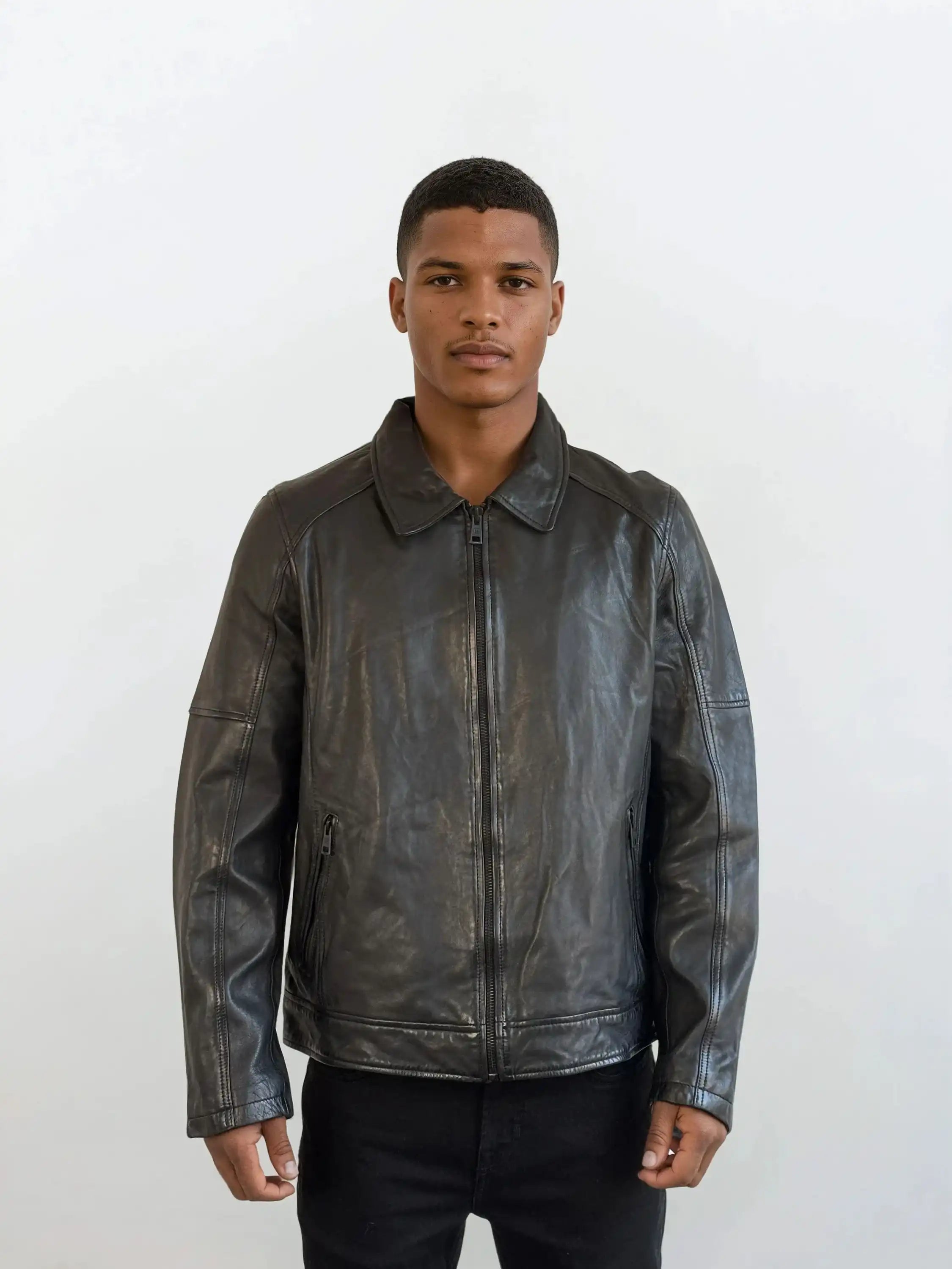 Vor weißem Hintergrund trägt eine Person die trueprodigy Herren Lederjacke aus Schafsleder in Schwarz mit Ärmel-Reißverschlüssen über schwarzem Shirt und Hose und blickt neutral in die Kamera.