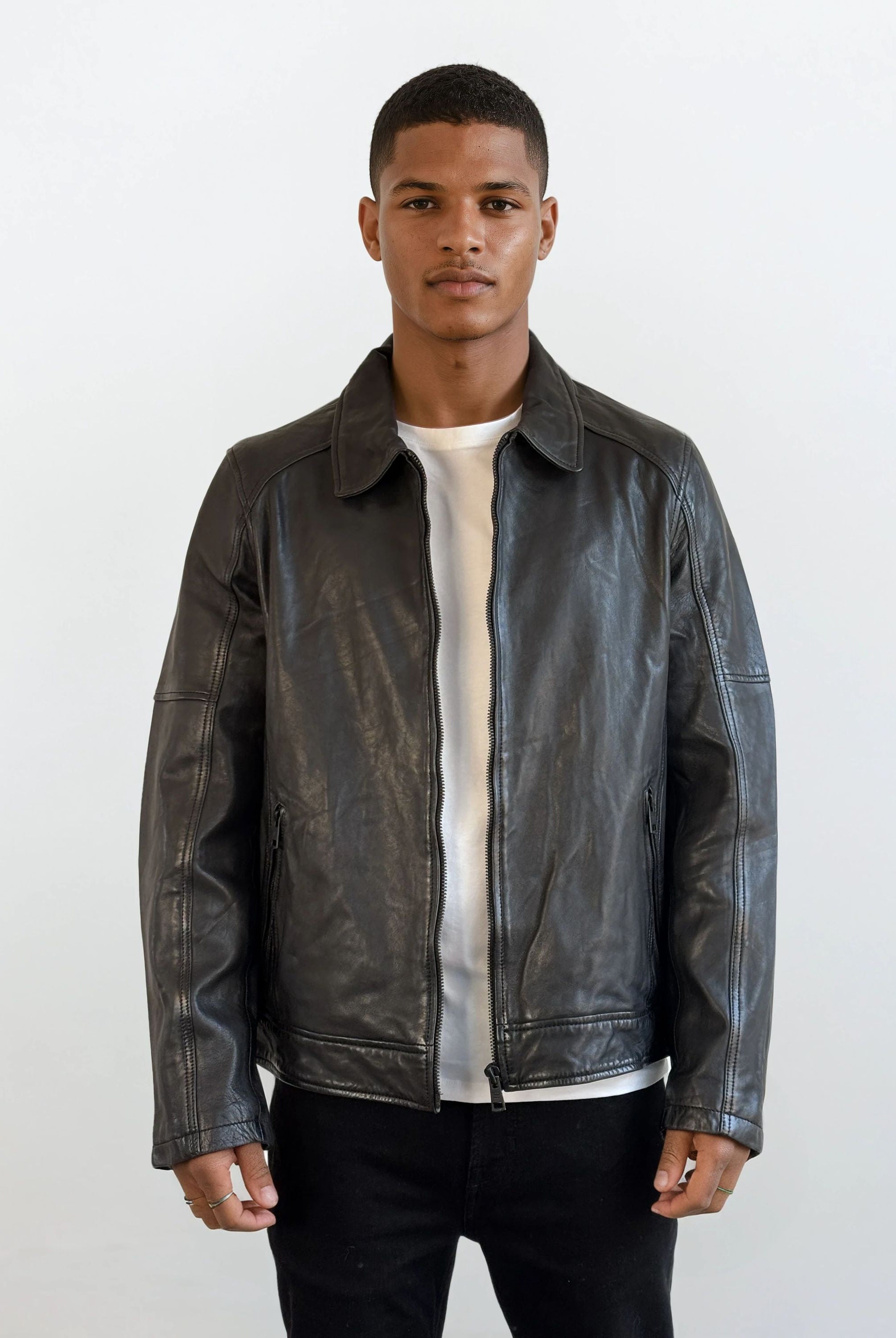 Ein junger Mann steht mit dem Gesicht nach vorne und trägt eine trueprodigy Herren Lederjacke aus Schafsleder in Schwarz mit Ärmel-Reißverschlüssen über einem weißen T-Shirt und einer schwarzen Hose vor einem schlichten weißen Hintergrund.