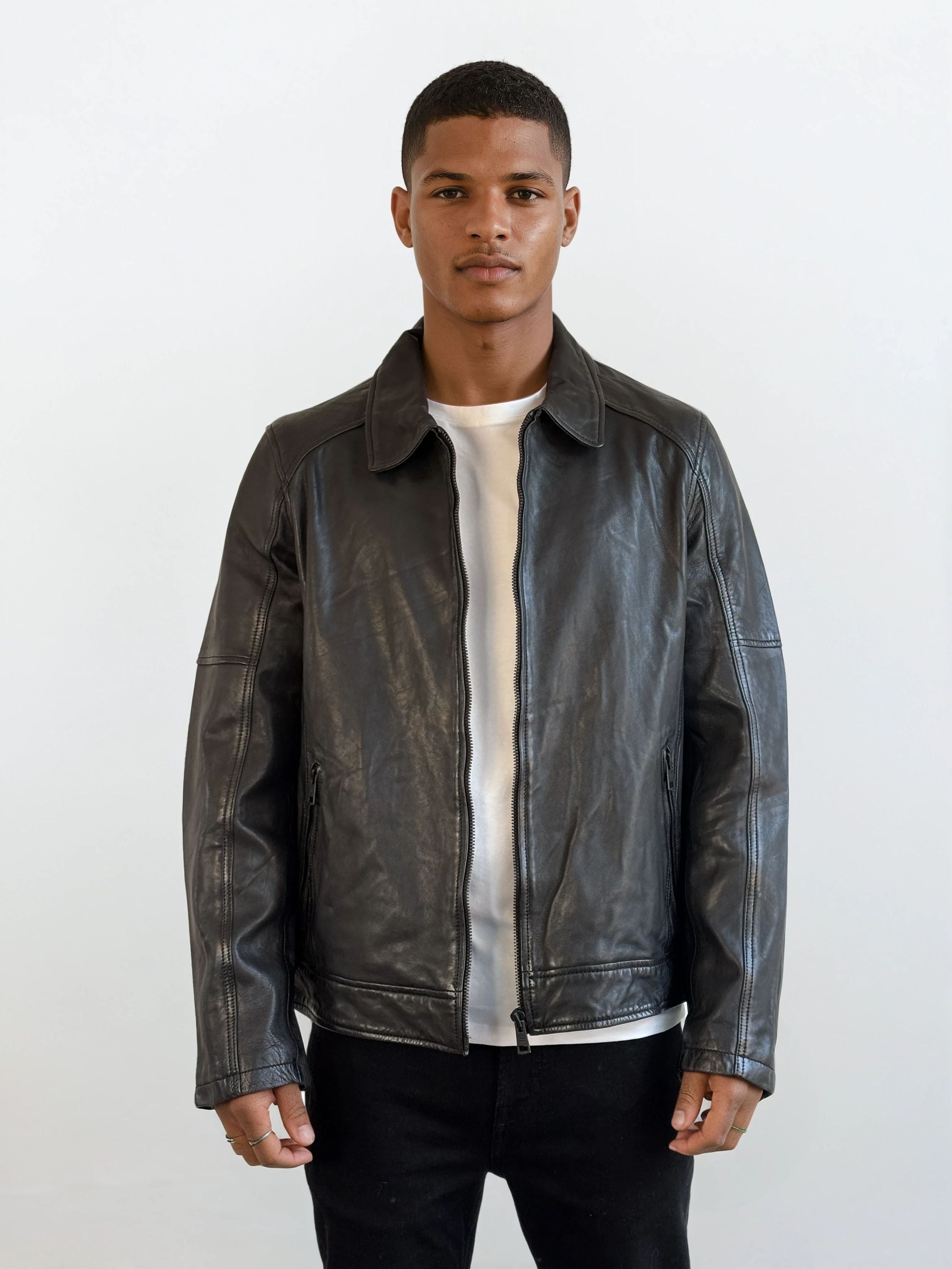 Ein junger Mann steht mit dem Gesicht nach vorne und trägt eine trueprodigy Herren Lederjacke aus Schafsleder in Schwarz mit Ärmel-Reißverschlüssen über einem weißen T-Shirt und einer schwarzen Hose vor einem schlichten weißen Hintergrund.