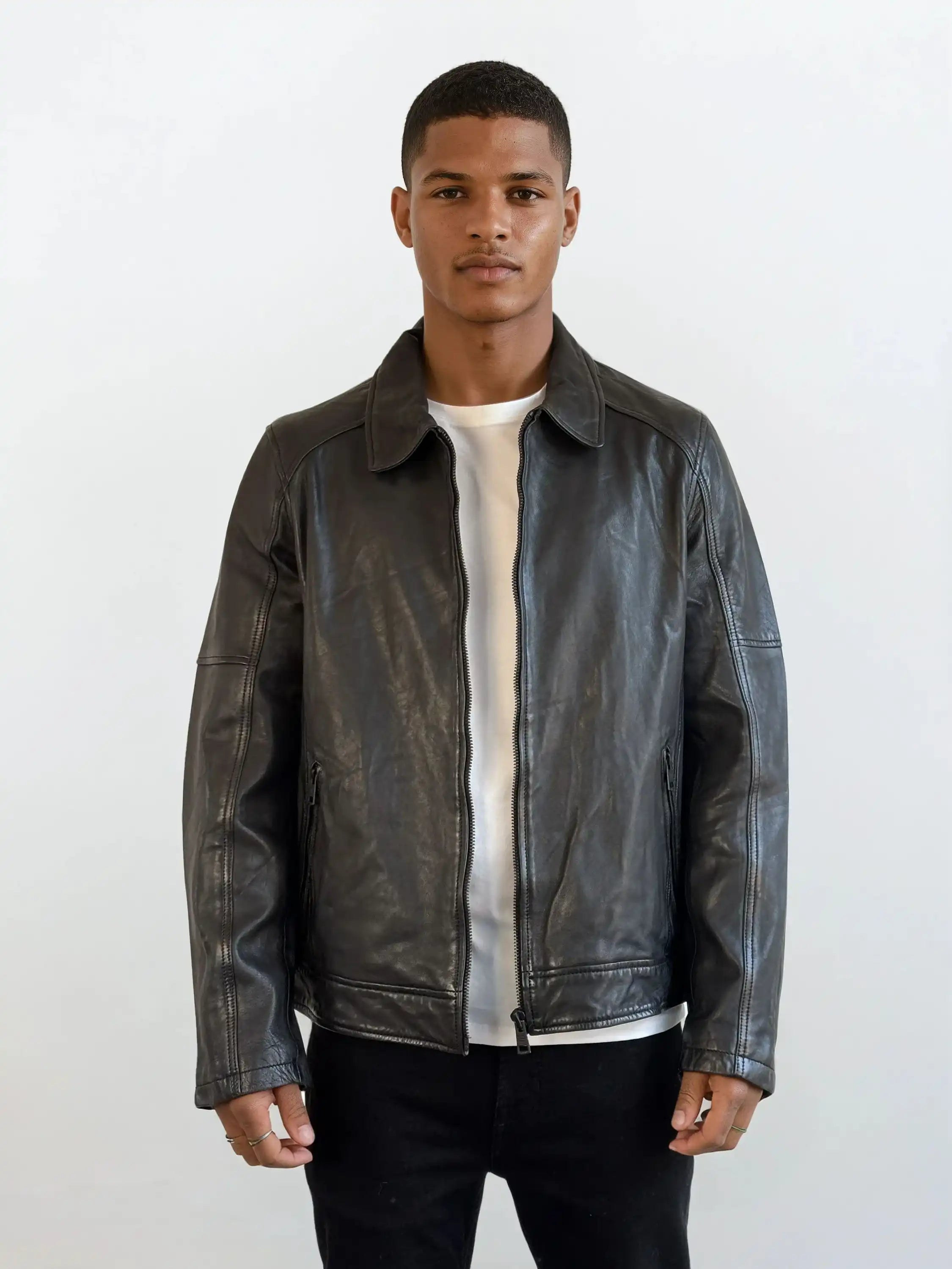 Ein junger Mann steht mit dem Gesicht nach vorne und trägt eine trueprodigy Herren Lederjacke aus Schafsleder in Schwarz mit Ärmel-Reißverschlüssen über einem weißen T-Shirt und einer schwarzen Hose vor einem schlichten weißen Hintergrund.