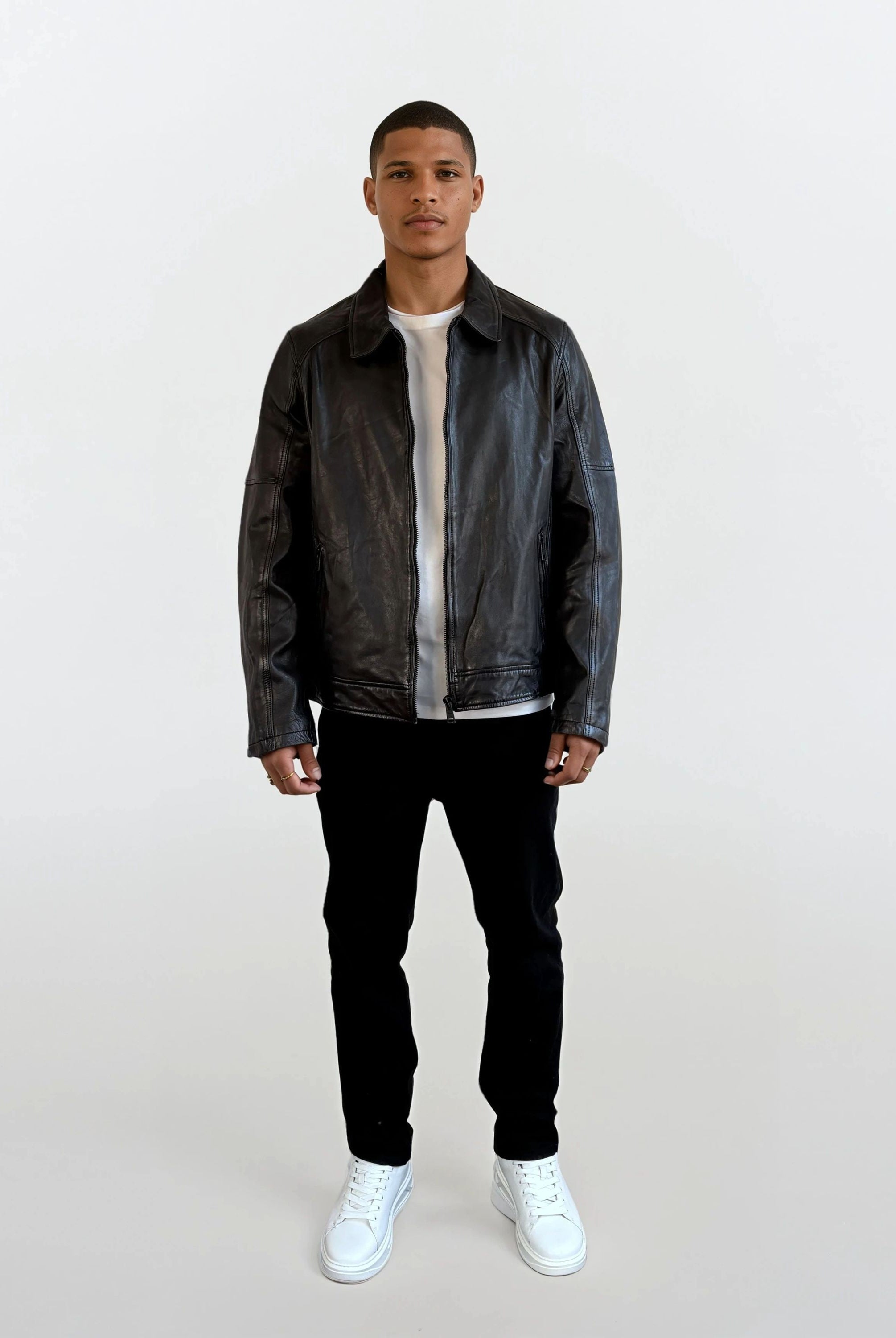 Eine Person steht vor weißem Hintergrund und trägt eine schwarze Herren Lederjacke aus Schafsleder mit Ärmel-Reißverschlüssen von trueprodigy, dazu ein weißes T-Shirt, schwarze Hosen und weiße Sneakers.