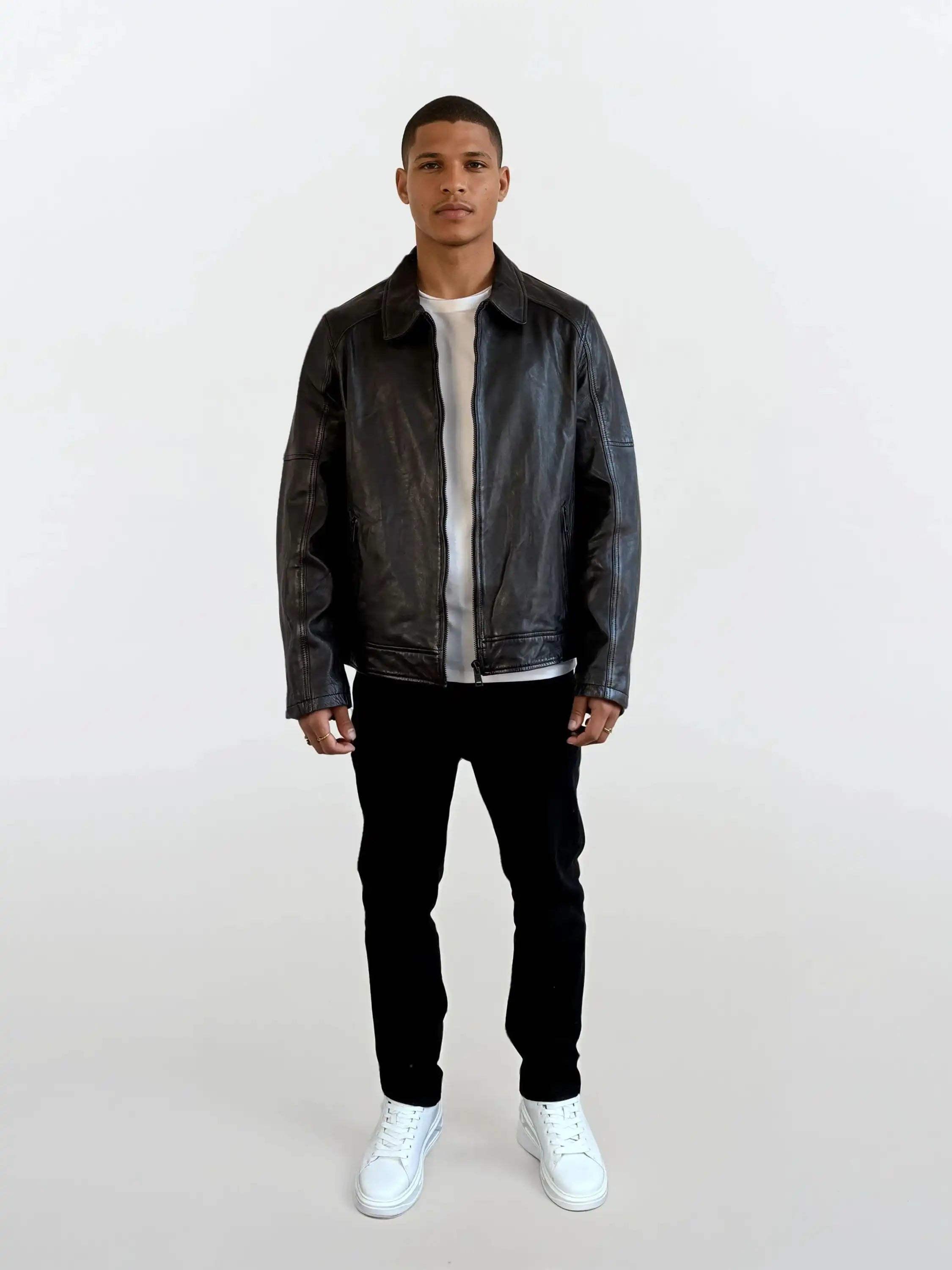 Eine Person steht vor weißem Hintergrund und trägt eine schwarze Herren Lederjacke aus Schafsleder mit Ärmel-Reißverschlüssen von trueprodigy, dazu ein weißes T-Shirt, schwarze Hosen und weiße Sneakers.