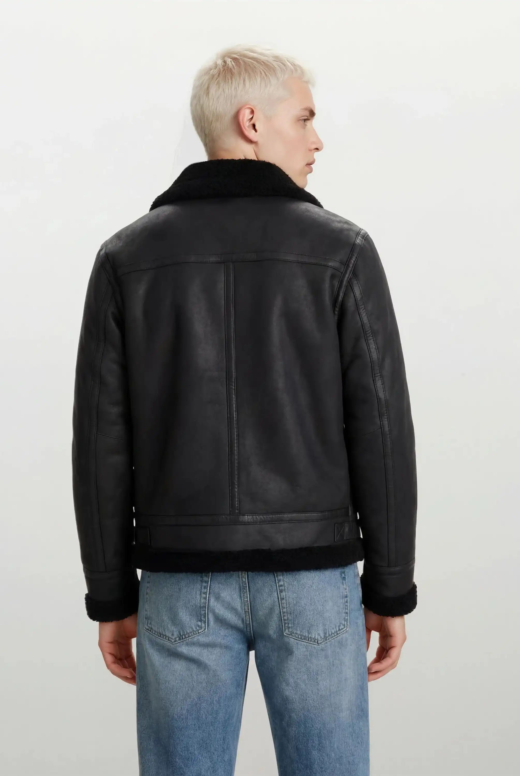 Man sieht eine Person mit kurzen, hellblonden Haaren von hinten, die eine trueprodigy Herren Lederjacke aus Schafsleder in Schwarz im Aviator-Stil mit Fellbesatz und hellblaue Jeans vor einem schlichten hellen Hintergrund trägt.