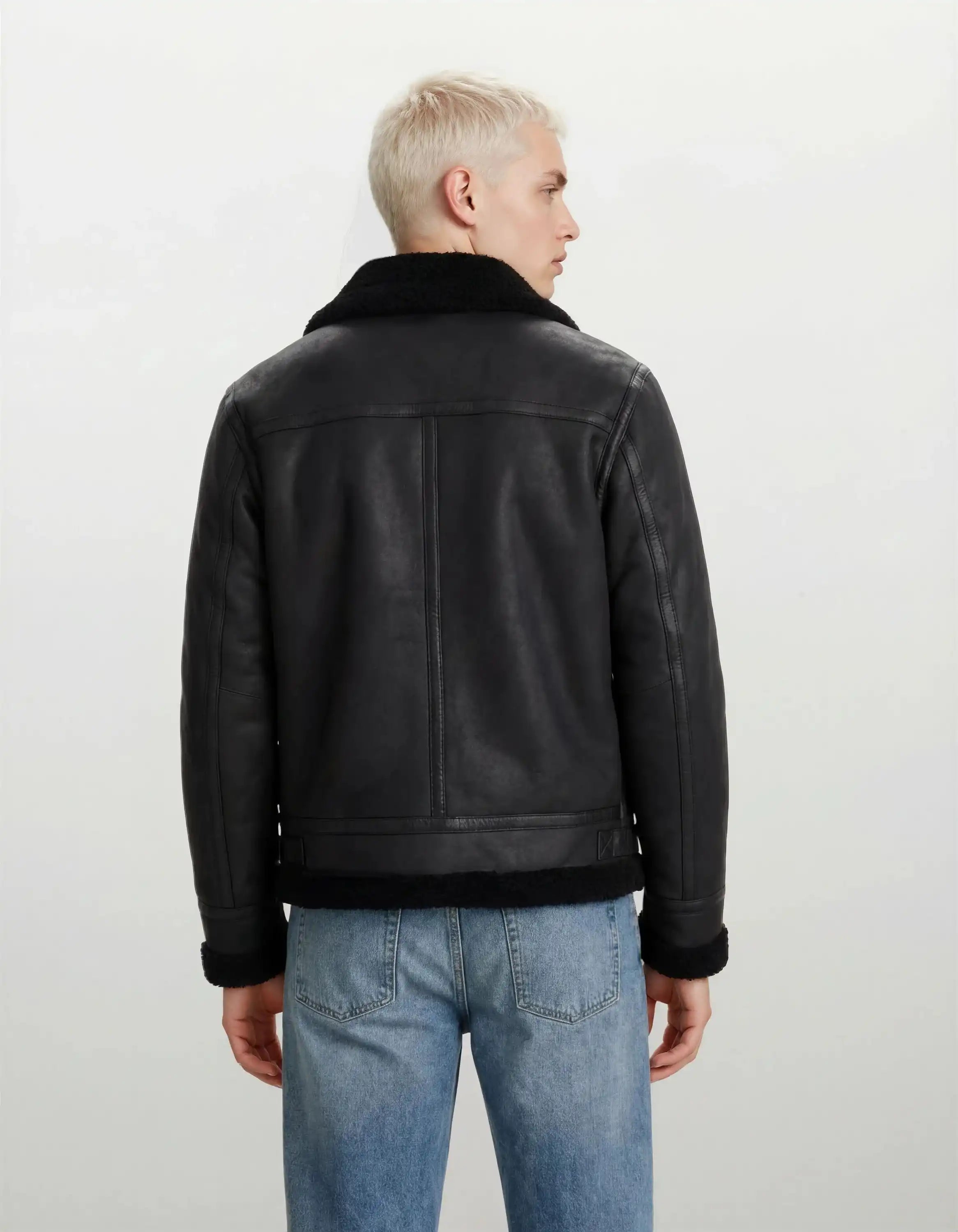 Man sieht eine Person mit kurzen, hellblonden Haaren von hinten, die eine trueprodigy Herren Lederjacke aus Schafsleder in Schwarz im Aviator-Stil mit Fellbesatz und hellblaue Jeans vor einem schlichten hellen Hintergrund trägt.