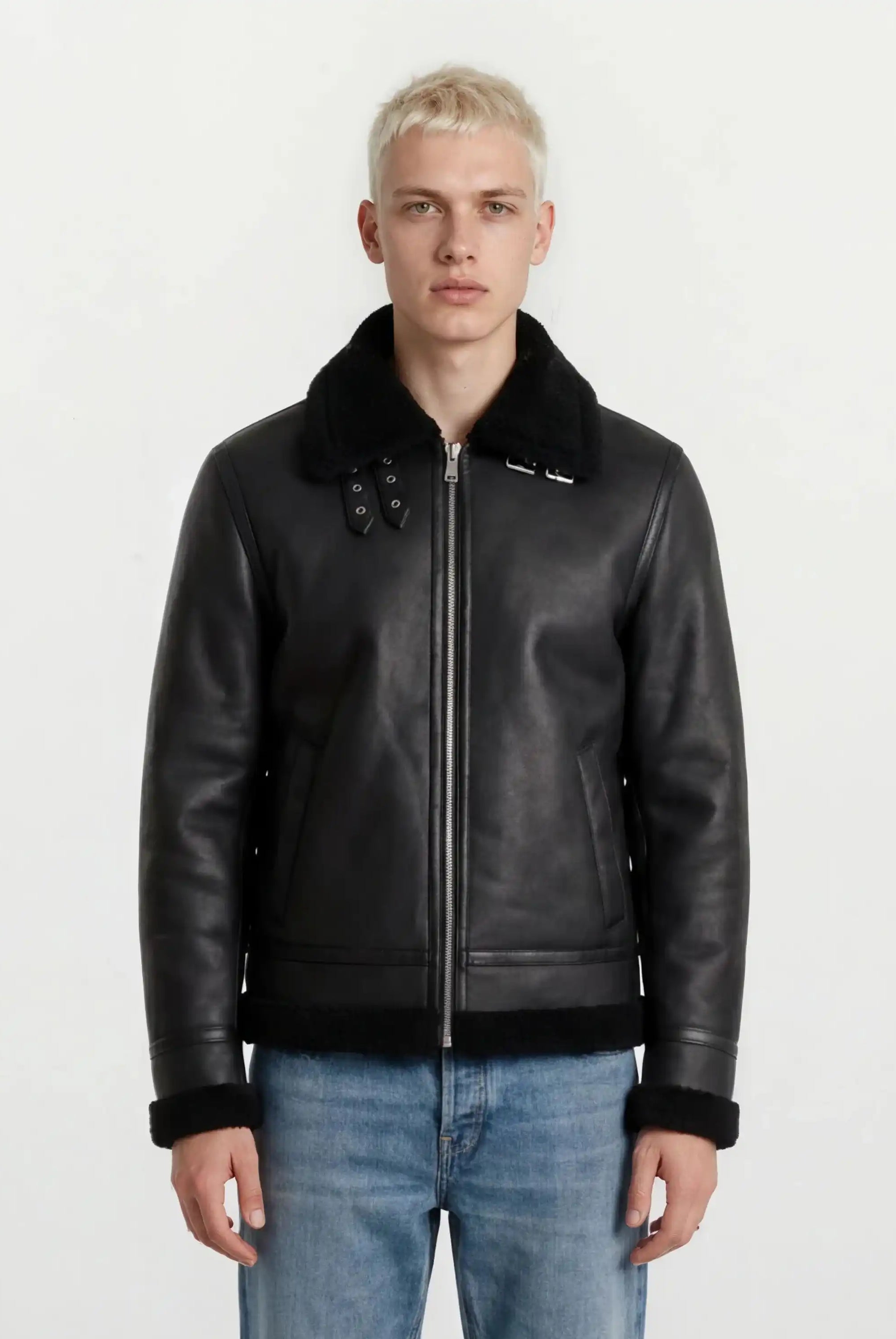 Eine Person mit kurzen blonden Haaren trägt die trueprodigy Herren Lederjacke aus Schafsleder in Schwarz im Aviator-Stil mit Fellbesatz und blaue Jeans, stehend vor einem schlichten weißen Hintergrund.