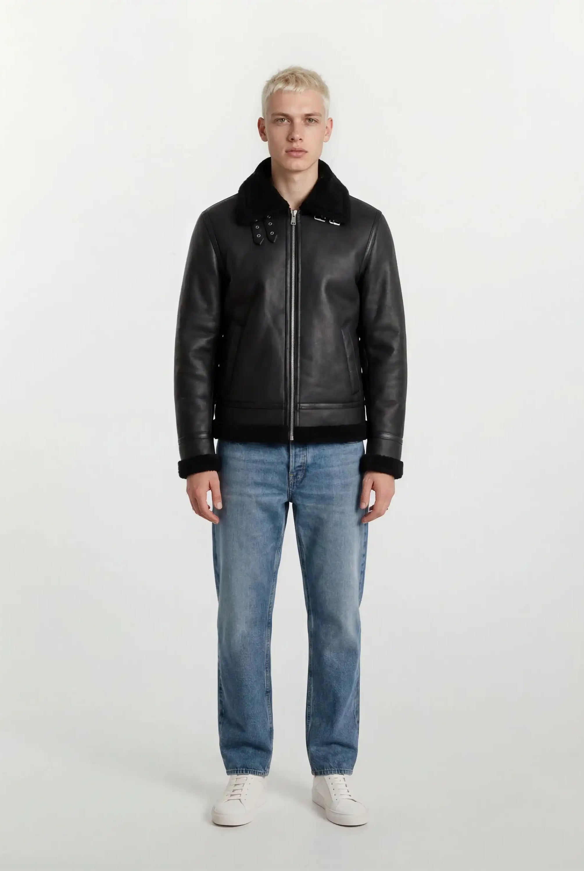 Eine Person mit kurzen blonden Haaren schaut nach vorne und trägt die trueprodigy Herren Lederjacke aus Schafsleder in Schwarz im Aviator-Stil mit Fellbesatz, gepaart mit hellblauen Jeans und weißen Turnschuhen vor einem schlichten hellen Hintergrund.