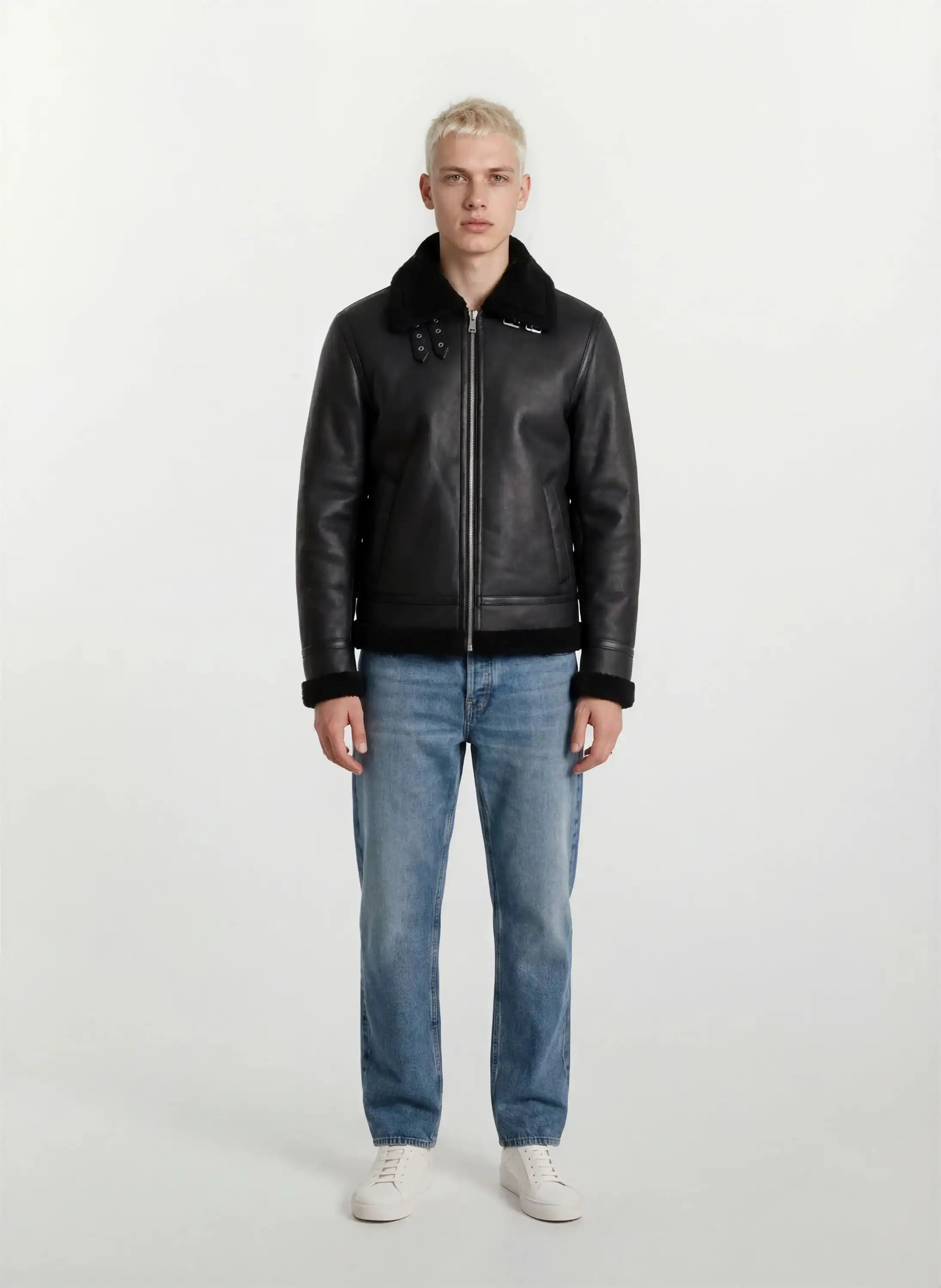 Eine Person mit kurzen blonden Haaren schaut nach vorne und trägt die trueprodigy Herren Lederjacke aus Schafsleder in Schwarz im Aviator-Stil mit Fellbesatz, gepaart mit hellblauen Jeans und weißen Turnschuhen vor einem schlichten hellen Hintergrund.