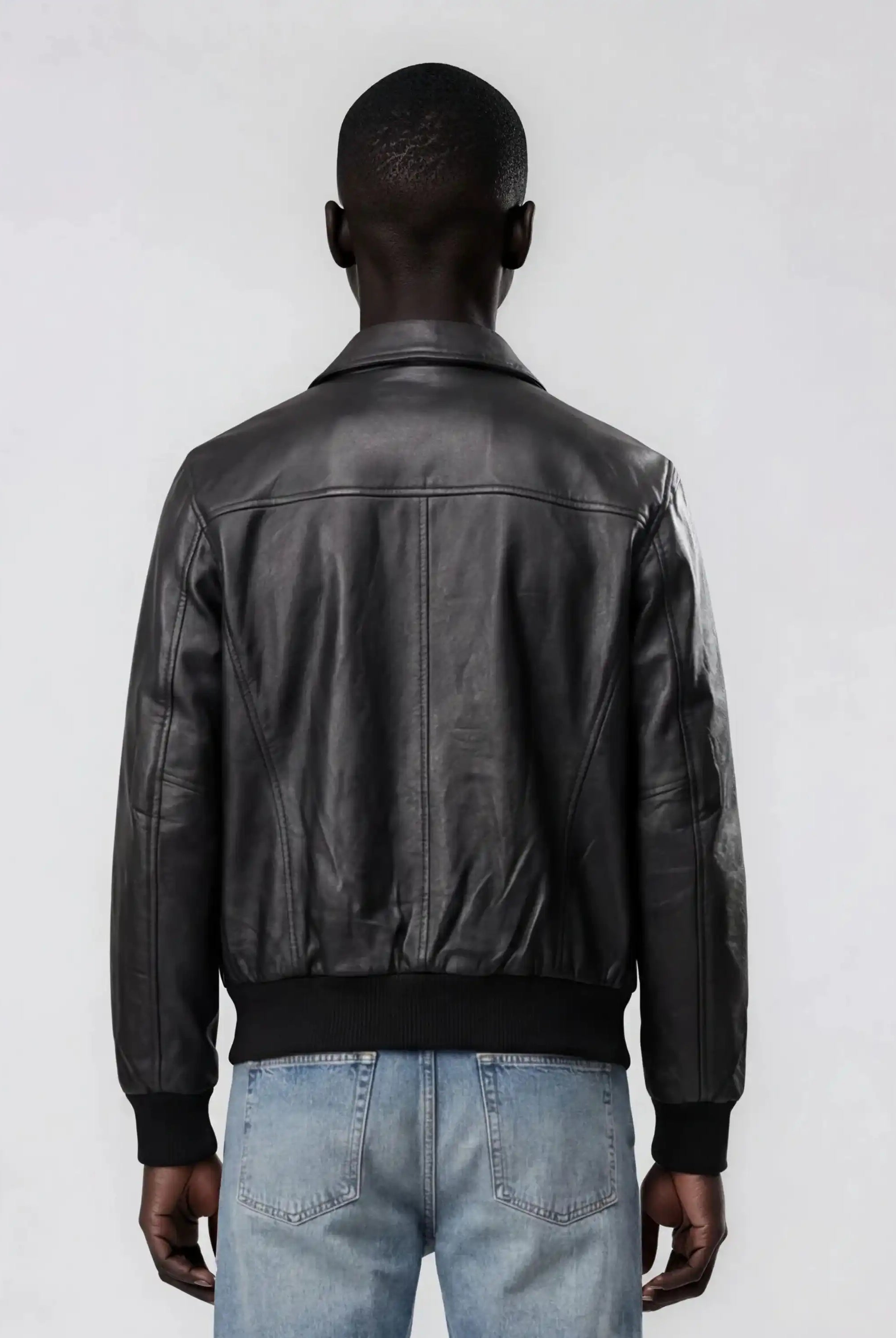 Man sieht eine Person von hinten, die die trueprodigy Herren Lederjacke aus Schafsleder in Schwarz mit klassischem Blouson-Schnitt und hellblaue Jeans vor einem schlichten, hellen Hintergrund trägt.