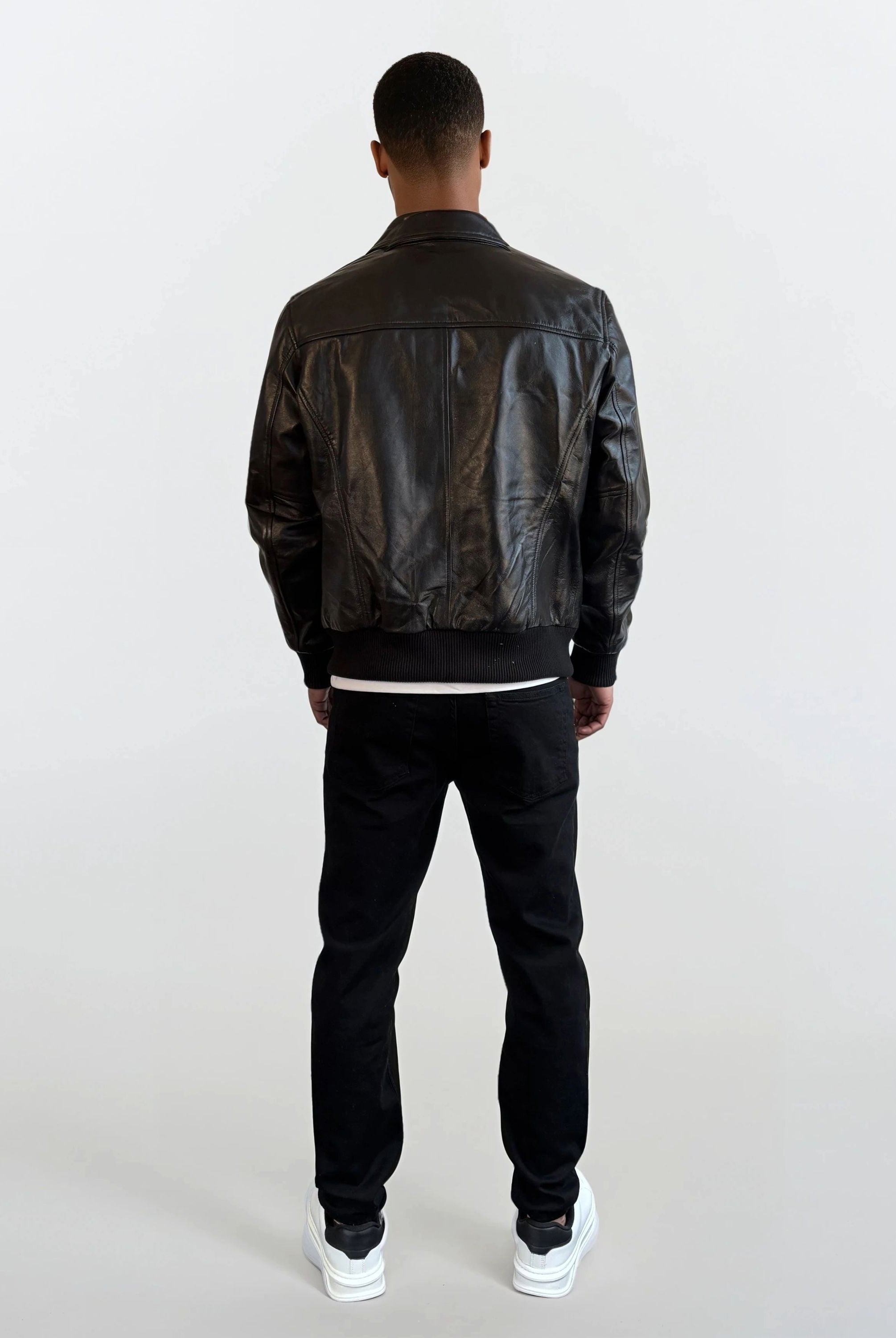 Eine Person trägt die trueprodigy Herren Lederjacke aus Schafsleder in Schwarz mit klassischem Blouson-Schnitt, kombiniert mit schwarzen Hosen und weißen Sneakern vor einem schlichten, hellen Hintergrund.