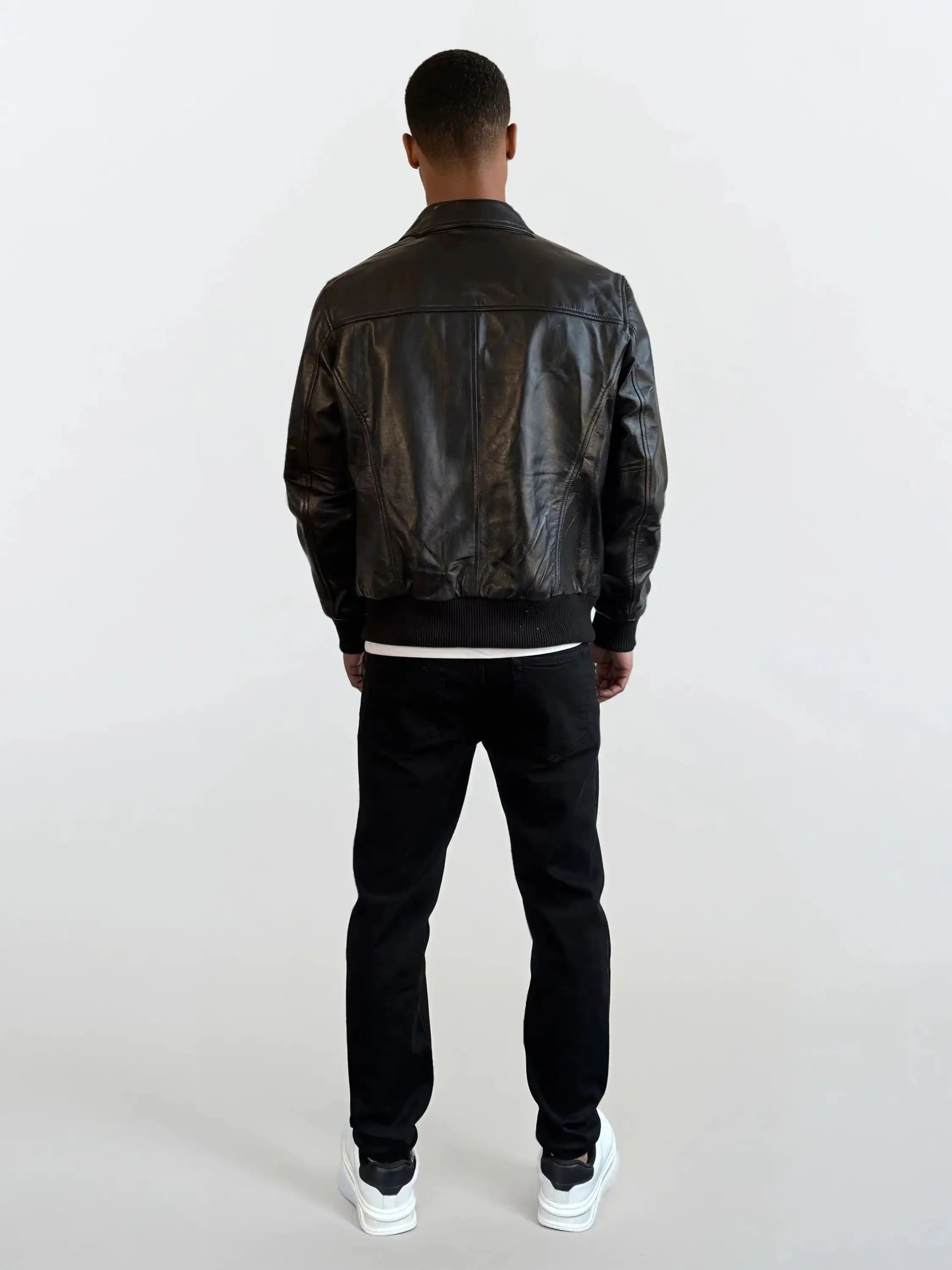 Eine Person trägt die trueprodigy Herren Lederjacke aus Schafsleder in Schwarz mit klassischem Blouson-Schnitt, kombiniert mit schwarzen Hosen und weißen Sneakern vor einem schlichten, hellen Hintergrund.