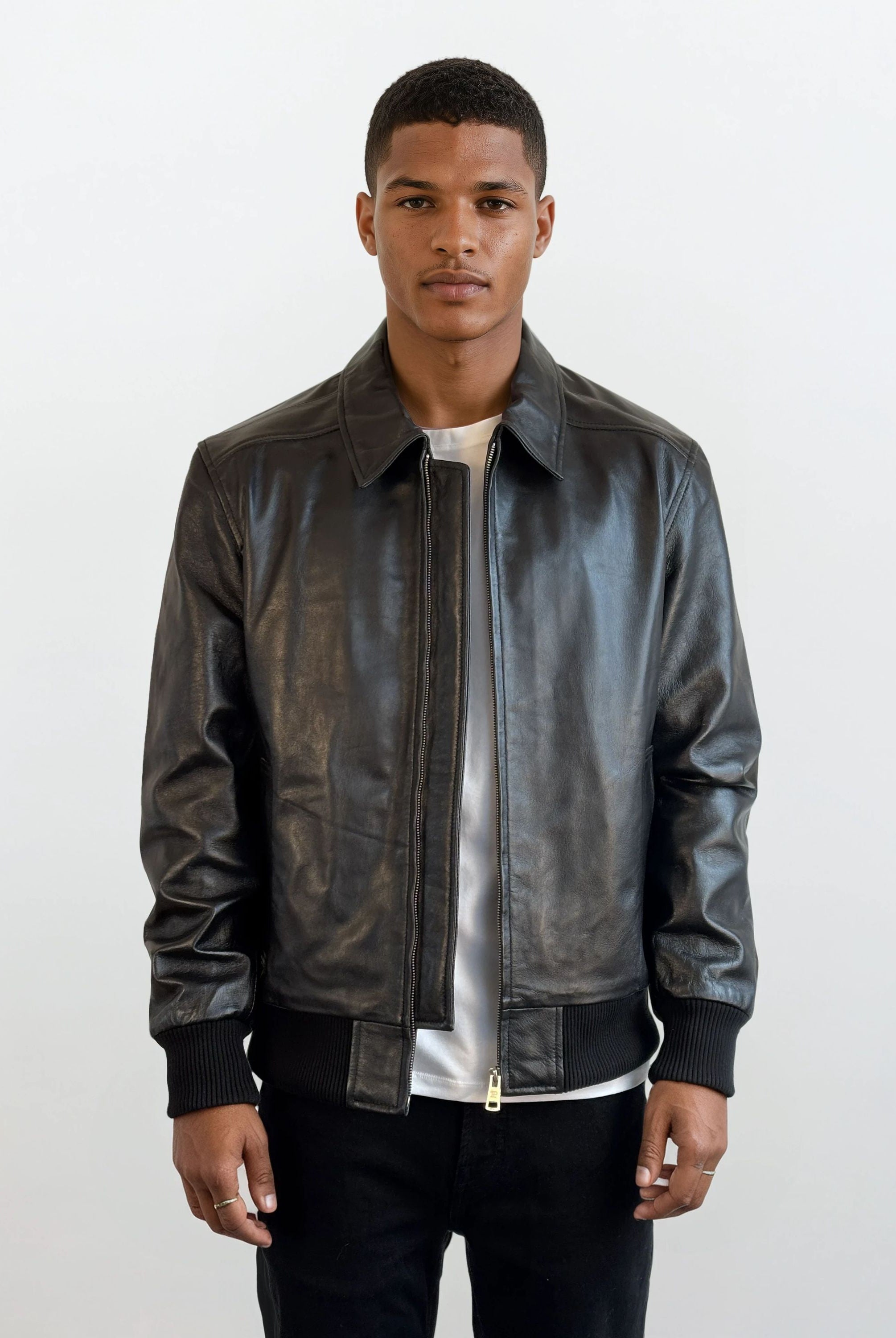 Ein Mann trägt die Herren Lederjacke aus Schafsleder in Schwarz mit klassischem Blouson-Schnitt von trueprodigy über einem weißen Shirt und schwarzen Hosen, steht entspannt vor hellem Hintergrund, die Hände an den Seiten.