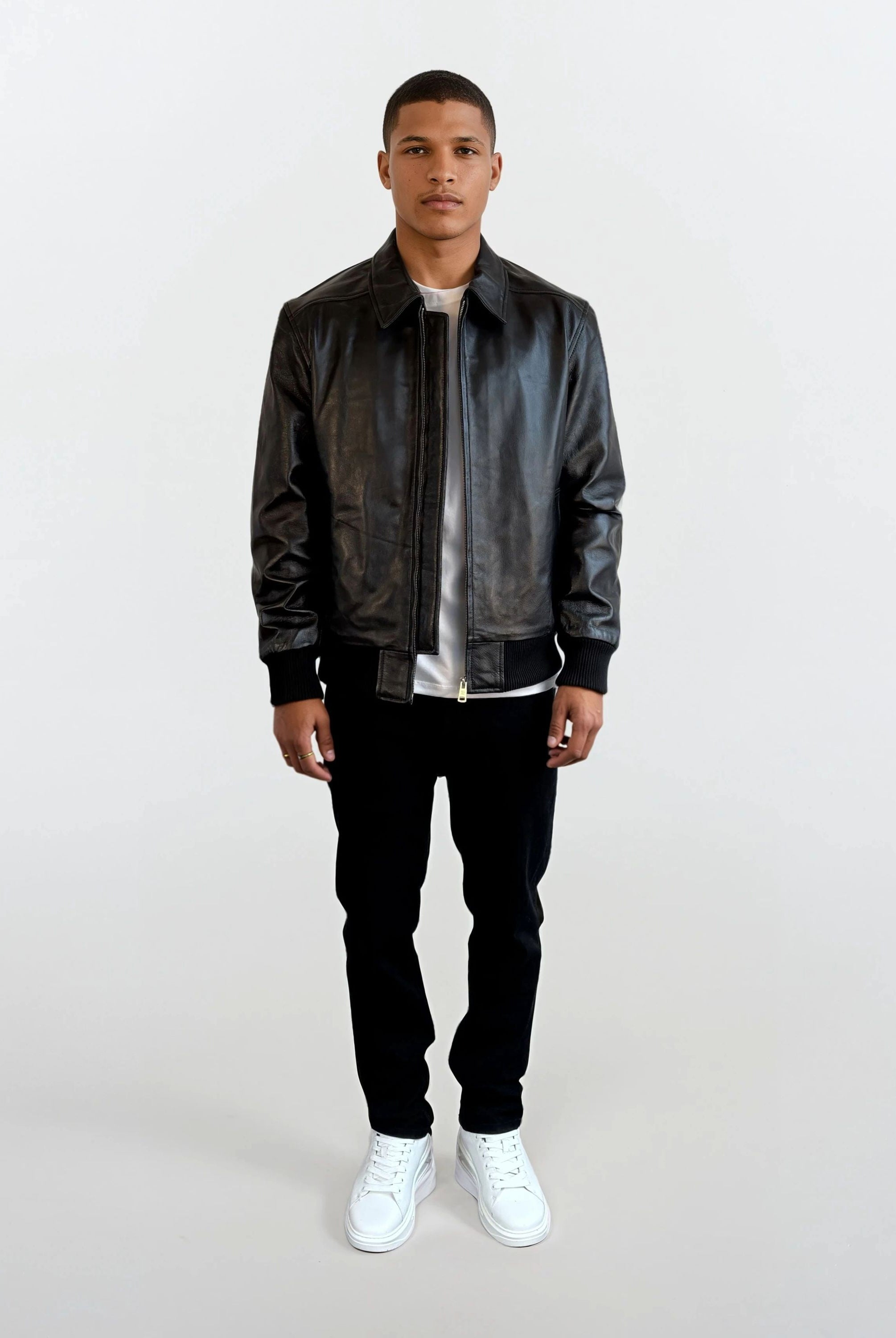 Eine Person trägt die schwarze Herren Lederjacke aus Schafsleder mit klassischem Blouson-Schnitt von trueprodigy, kombiniert mit weißem T-Shirt, schwarzer Hose und weißen Sneakers vor schlichtem weißem Hintergrund.