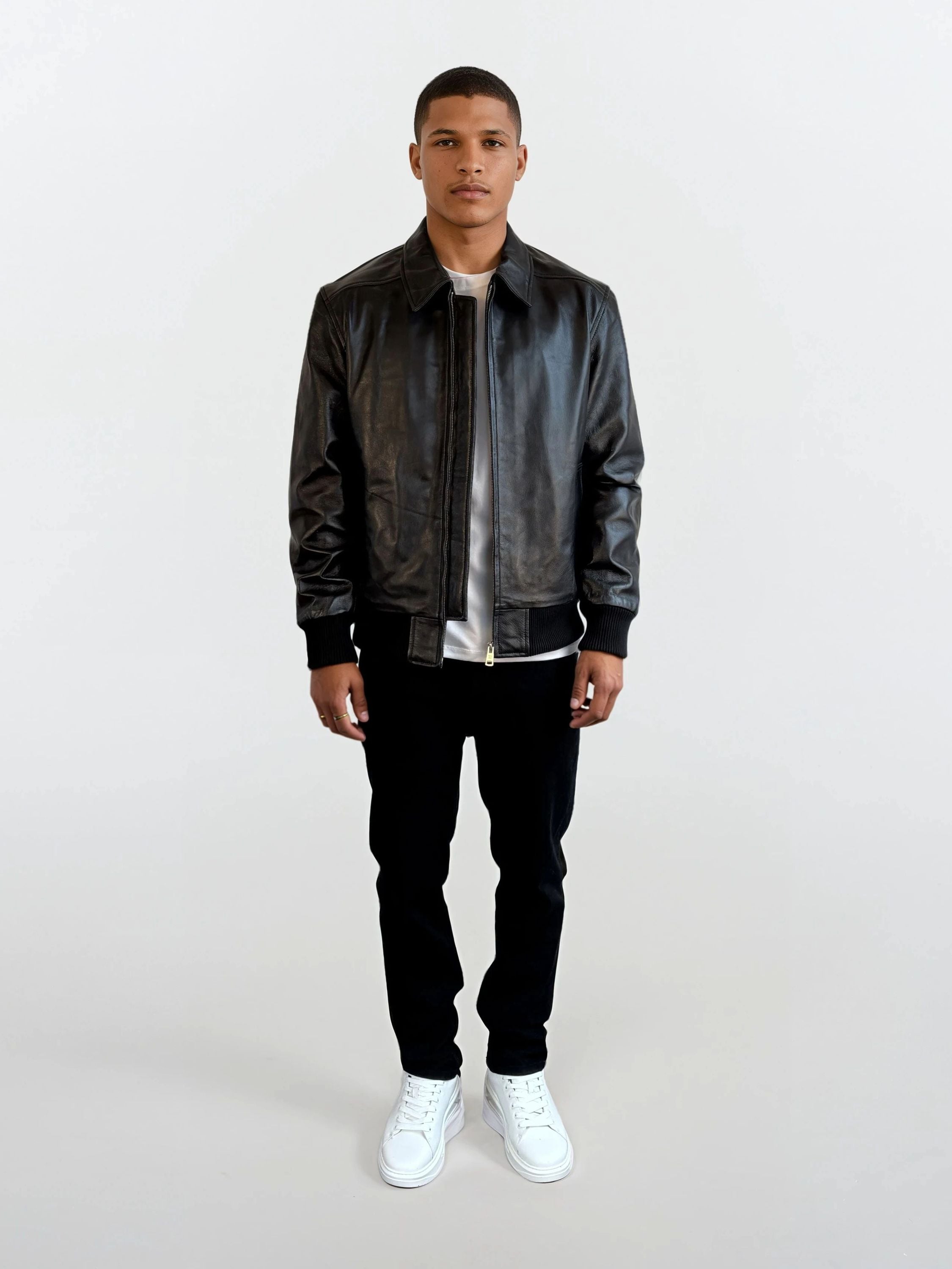 Eine Person trägt die schwarze Herren Lederjacke aus Schafsleder mit klassischem Blouson-Schnitt von trueprodigy, kombiniert mit weißem T-Shirt, schwarzer Hose und weißen Sneakers vor schlichtem weißem Hintergrund.