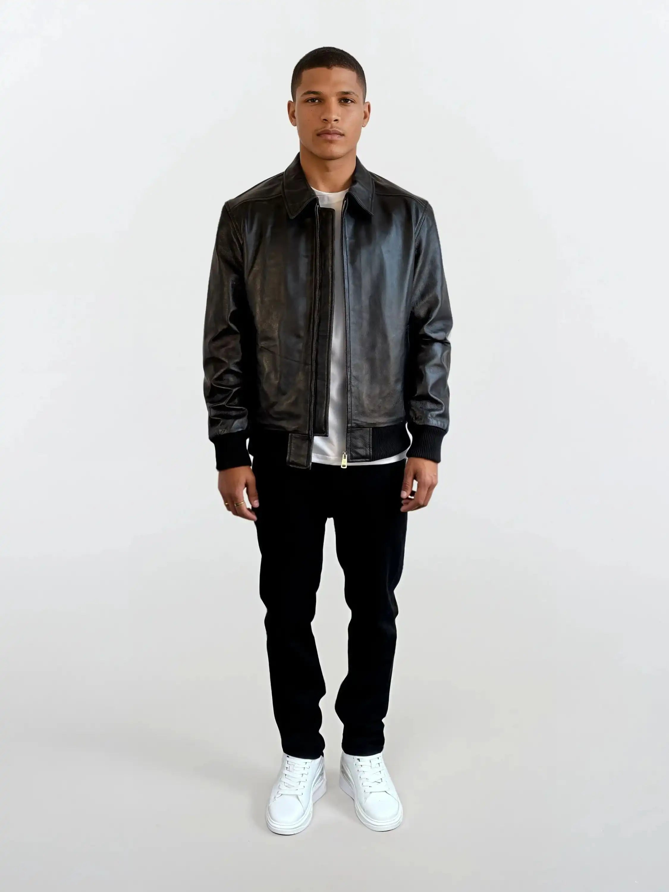 Eine Person trägt die schwarze Herren Lederjacke aus Schafsleder mit klassischem Blouson-Schnitt von trueprodigy, kombiniert mit weißem T-Shirt, schwarzer Hose und weißen Sneakers vor schlichtem weißem Hintergrund.