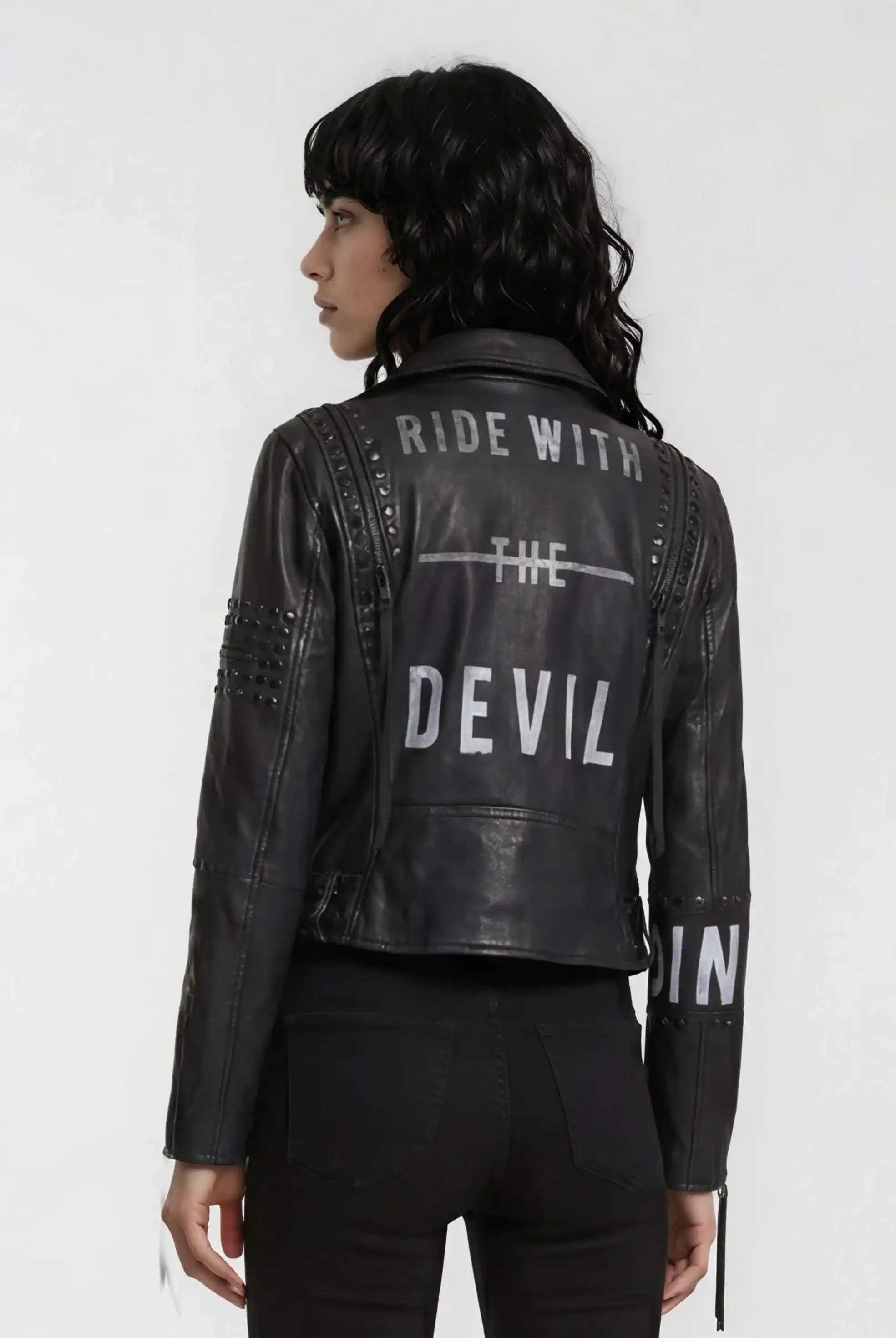 Eine Person mit langem, welligem schwarzem Haar trägt eine trueprodigy Bikerlederjacke mit silbernen Nieten, weißem "RIDE WITH THE DEVIL"-Aufdruck und dazu passenden schwarzen Hosen.
