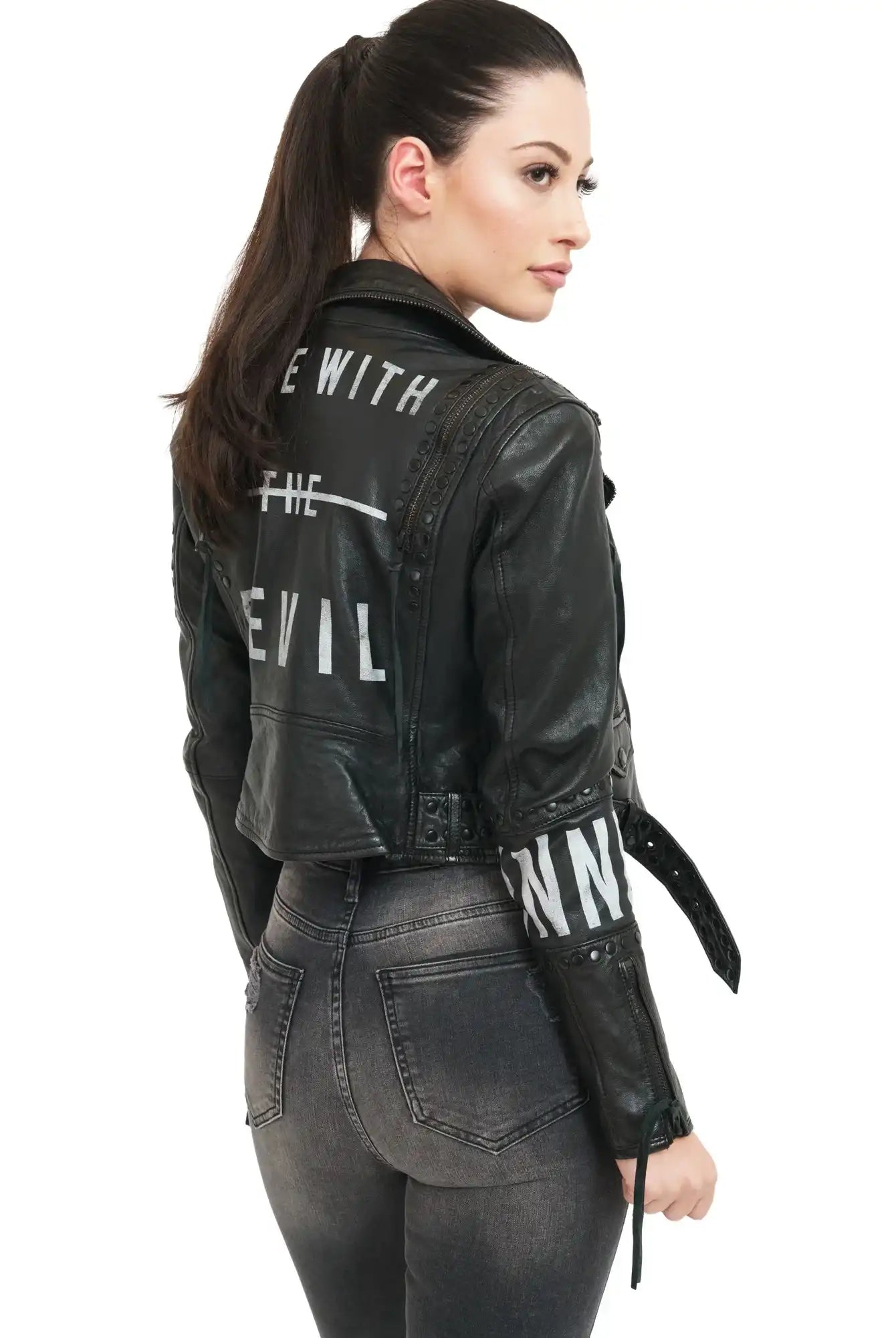 Eine Frau mit hochgestecktem Pferdeschwanz trägt die trueprodigy Ava Bikerlederjacke mit Nieten und Print, mit weißem "THE WITH THE DEVIL"-Schriftzug auf dem Rücken, und eine taillierte graue Jeans. Sie ist leicht zur Seite gedreht und schaut über ihre Schulter.