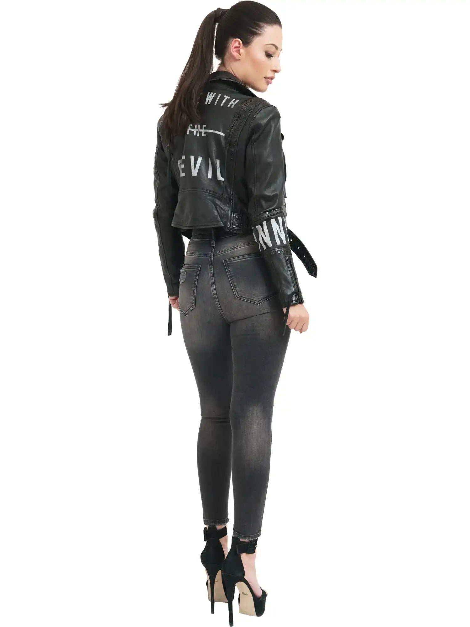 Eine Frau mit Pferdeschwanz steht mit dem Gesicht zur Wand. Sie trägt die trueprodigy Ava Bikerlederjacke mit Nieten und Print mit der Aufschrift WITH THE DEVIL und dunkelgraue Skinny-Jeans, ergänzt durch schwarze Stöckelschuhe vor weißem Hintergrund.