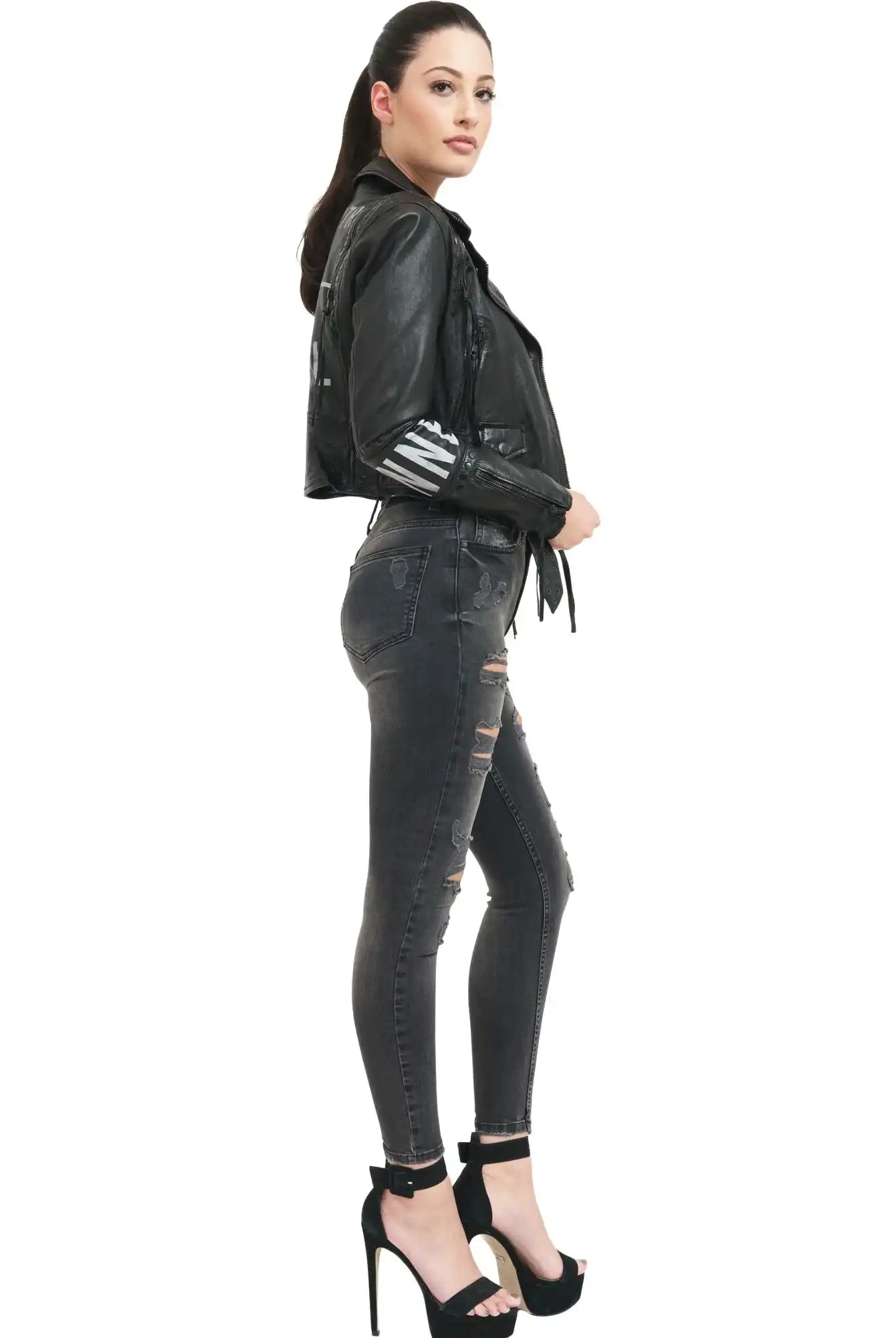 Eine Frau steht im Profil vor einem weißen Hintergrund. Sie trägt die trueprodigy Ava Bikerlederjacke mit Nieten und Print, dunkle zerrissene Skinny Jeans und schwarze hochhackige Sandalen.