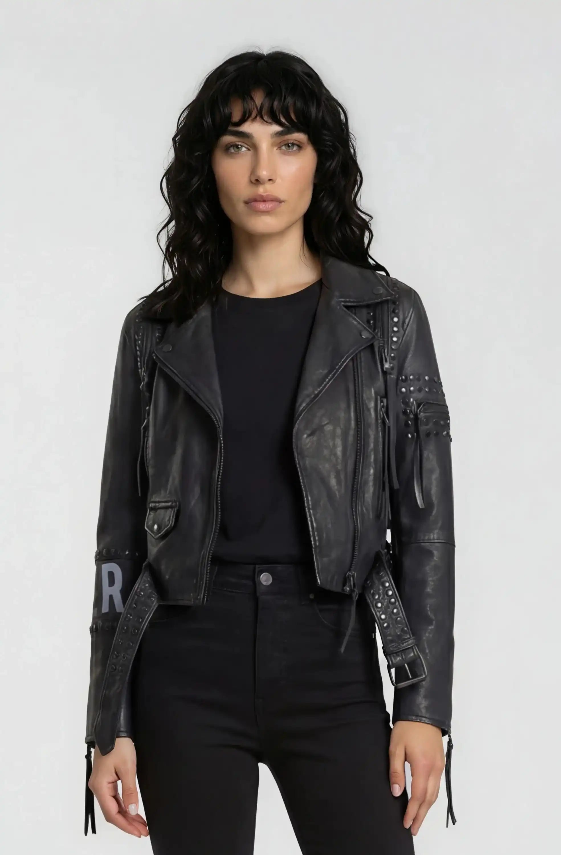 Eine Frau mit welligem, dunklem Haar trägt die Bikerlederjacke mit Nieten und Print von trueprodigy über schwarzem Shirt und Hose, stehend vor einem schlichten, hellgrauen Hintergrund.