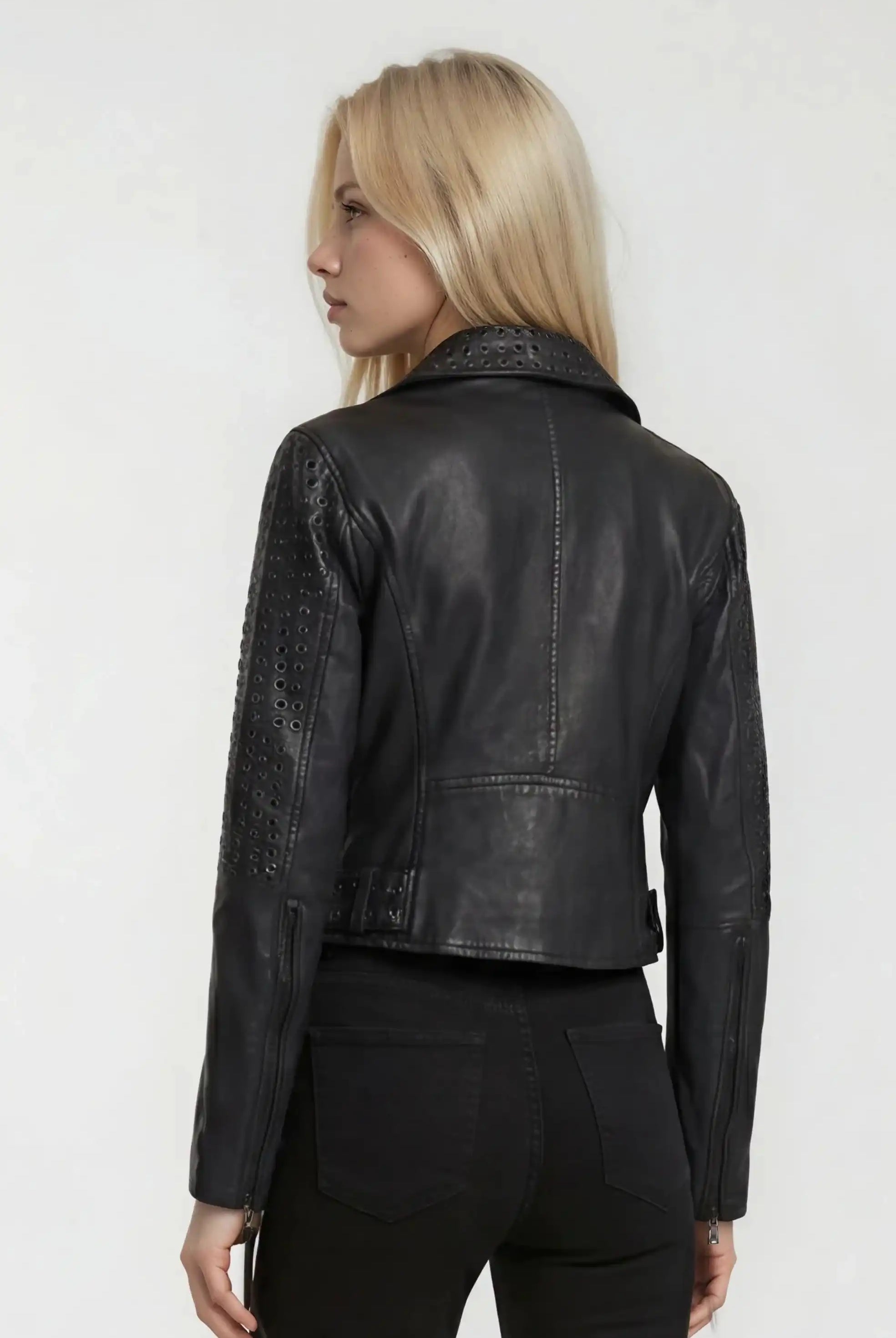 Eine Person mit langen blonden Haaren, bekleidet mit einer schwarzen Trueprodigy Bikerlederjacke mit Ösen und einer schwarzen Hose, steht von der Kamera abgewandt vor einem schlichten hellen Hintergrund.