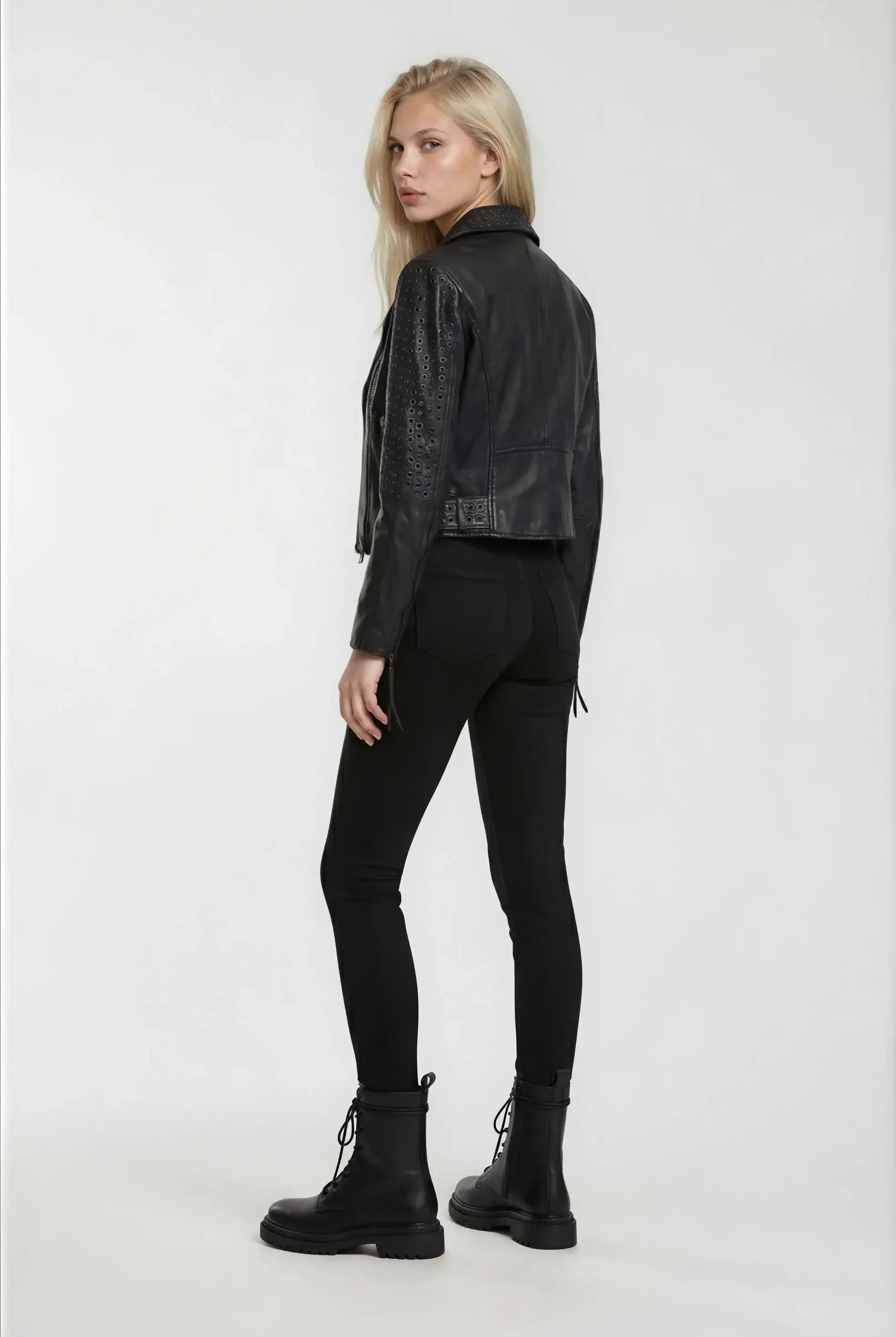Eine Frau mit langen blonden Haaren blickt über ihre Schulter. Sie trägt eine schwarze Trueprodigy Bikerlederjacke mit Ösen, eine schwarze Hose und schwarze Schnürstiefel vor einem schlichten weißen Hintergrund.