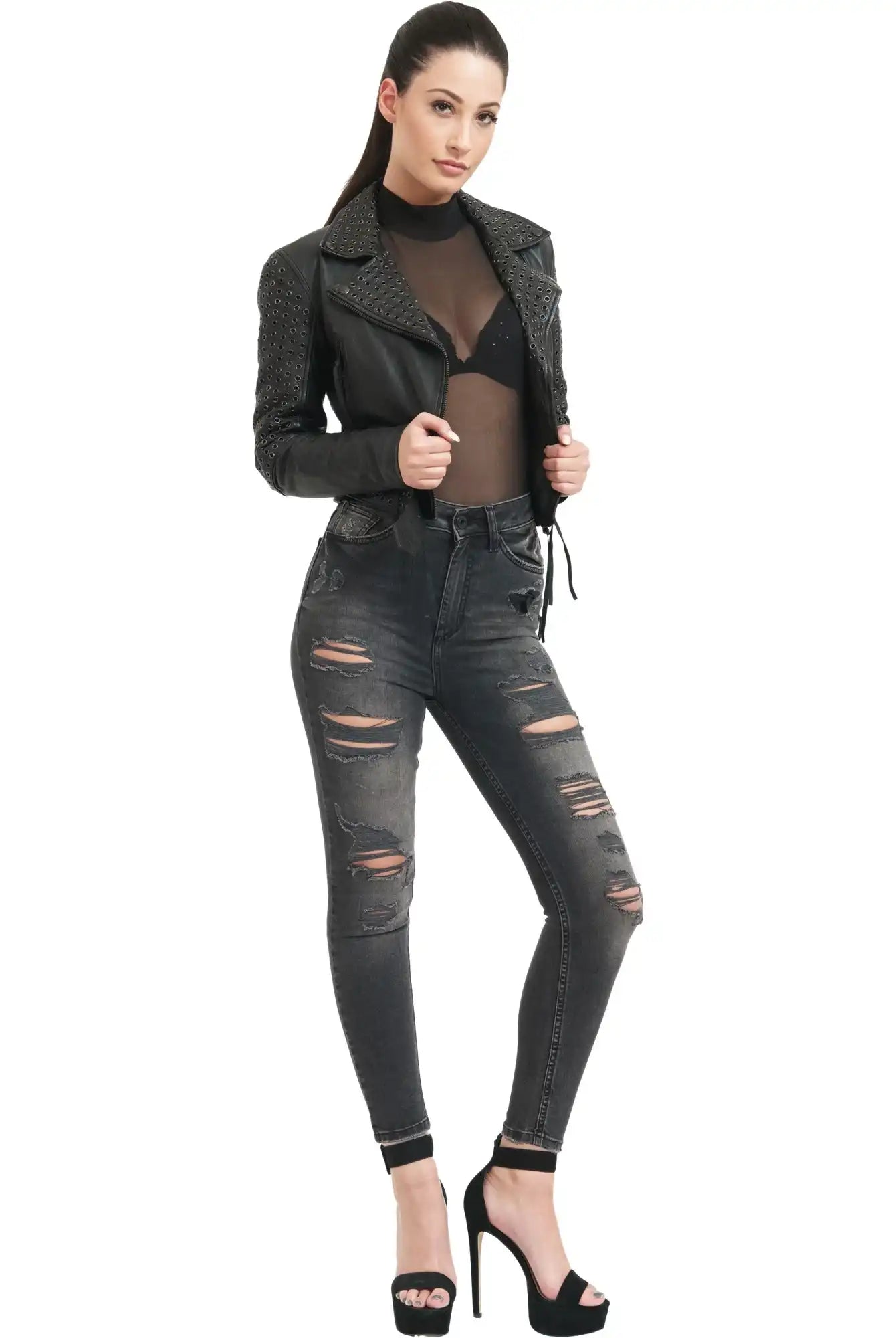 Eine Frau steht vor einem weißen Hintergrund und trägt eine schwarze Trueprodigy Raven Bikerlederjacke mit Ösen, ein durchsichtiges schwarzes Oberteil mit sichtbarem schwarzem BH, ausgebeulte Skinny Jeans und schwarze hochhackige Sandalen.