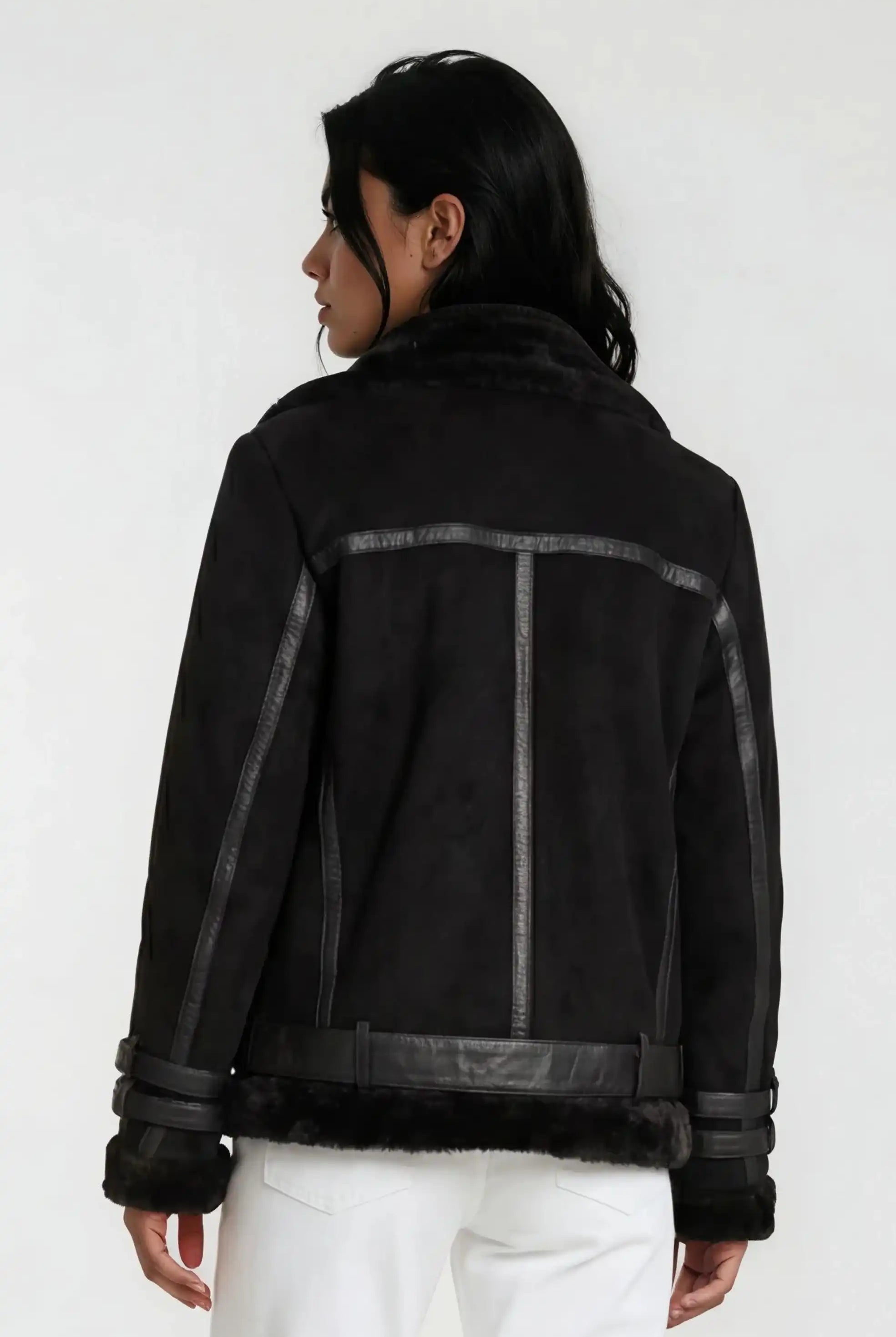 Eine Person mit langen dunklen Haaren trägt eine schwarze Trueprodigy Wilderlederjacke mit Gürtel und eine weiße Hose und blickt von der Kamera weg vor einem schlichten hellen Hintergrund.