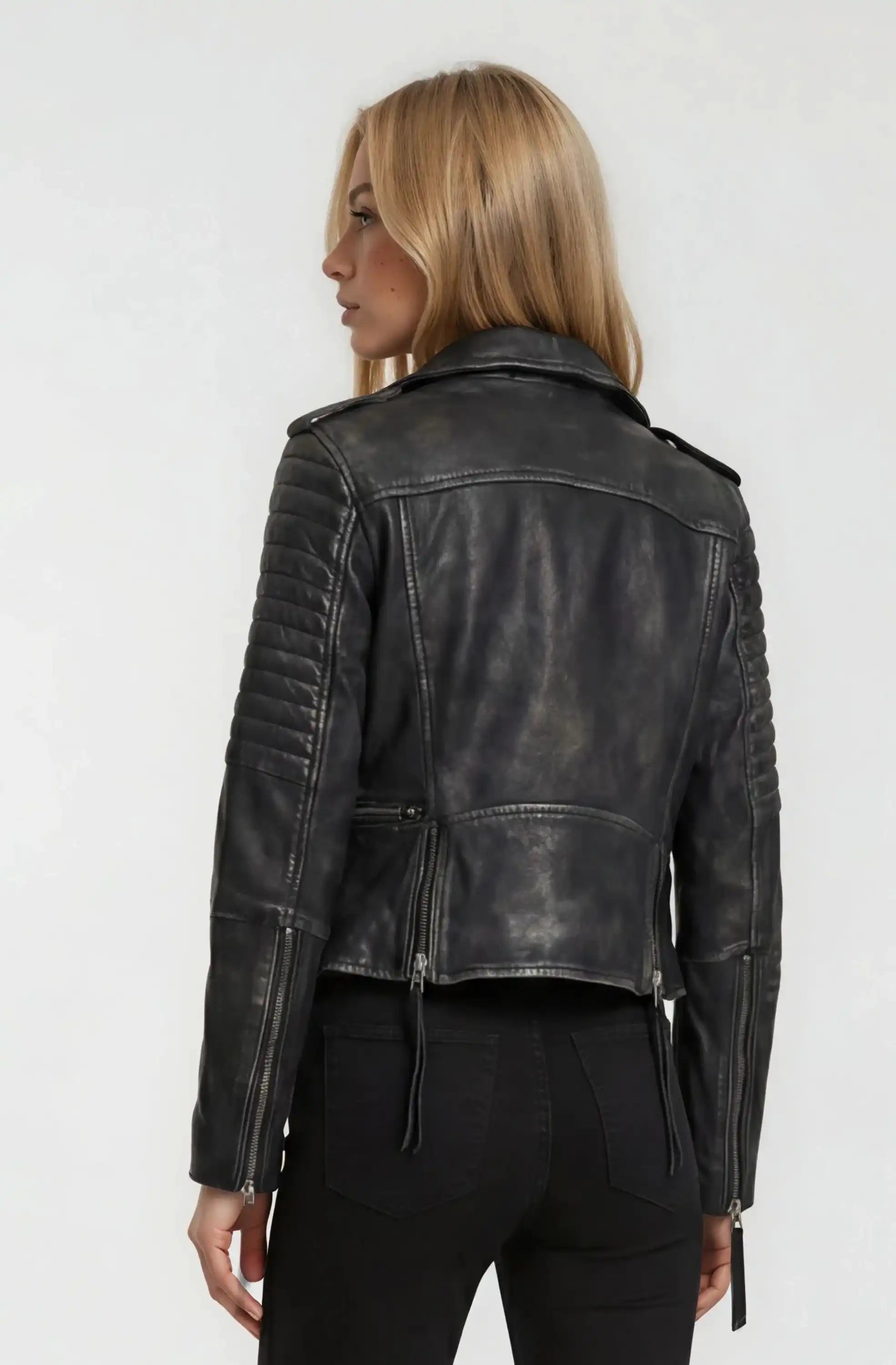 Eine Person mit langem, glattem blondem Haar wird von hinten gezeigt und trägt eine schwarze Bikerlederjacke Vintage Optik von trueprodigy und eine schwarze Jeans vor einem schlichten, hellen Hintergrund.