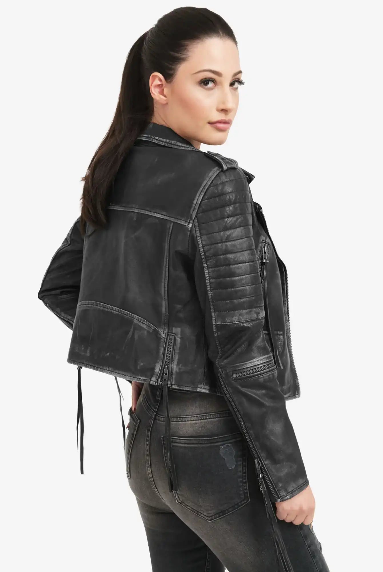 Eine Frau mit langen dunklen Haaren in einem Pferdeschwanz modelliert die trueprodigy Avril Bikerlederjacke Vintage Optik, gepaart mit einer dunklen Jeans. Sie steht schräg zur Kamera und blickt über ihre Schulter vor einem schlichten weißen Hintergrund.