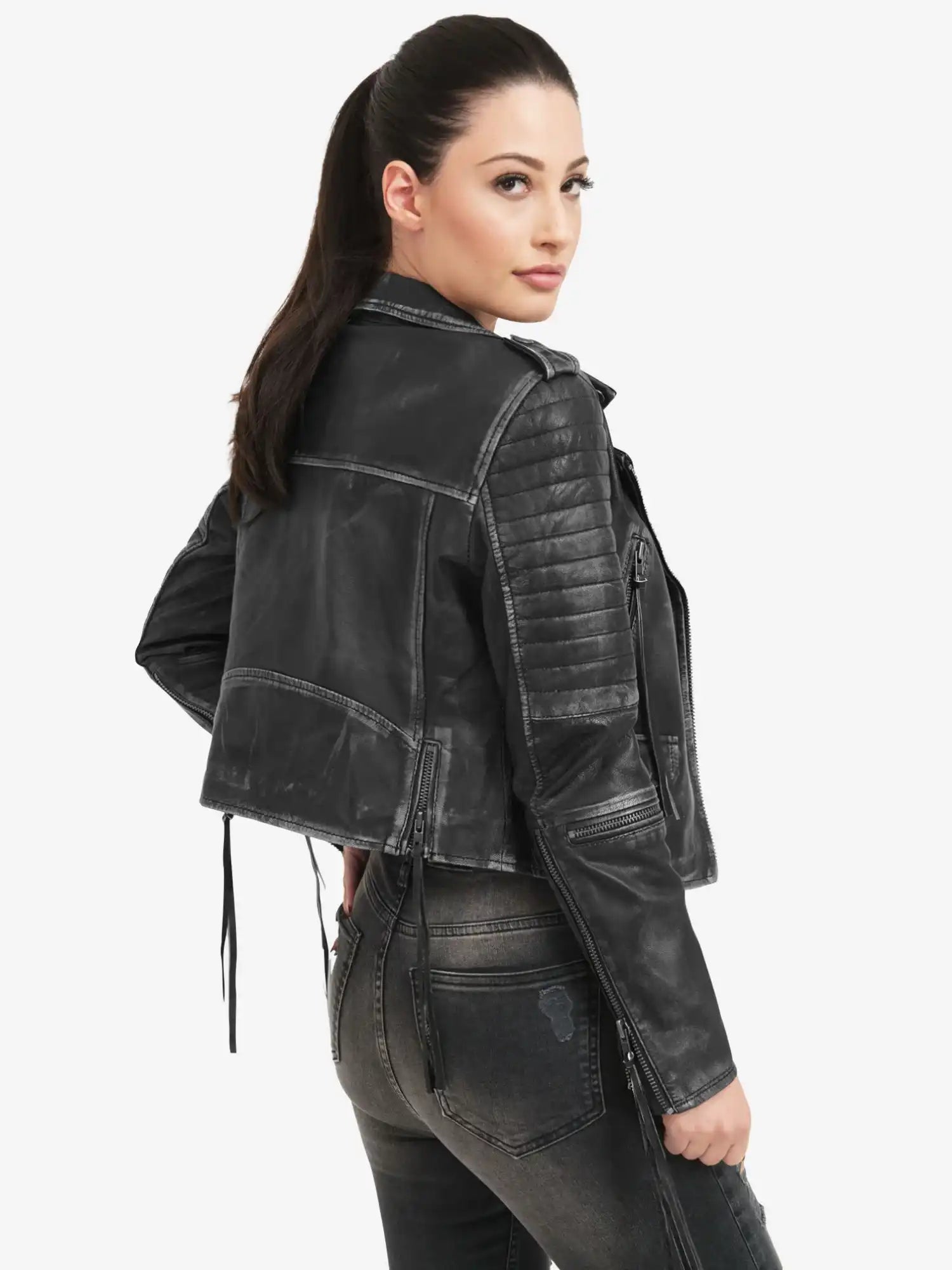 Eine Frau mit langen dunklen Haaren in einem Pferdeschwanz modelliert die trueprodigy Avril Bikerlederjacke Vintage Optik, gepaart mit einer dunklen Jeans. Sie steht schräg zur Kamera und blickt über ihre Schulter vor einem schlichten weißen Hintergrund.