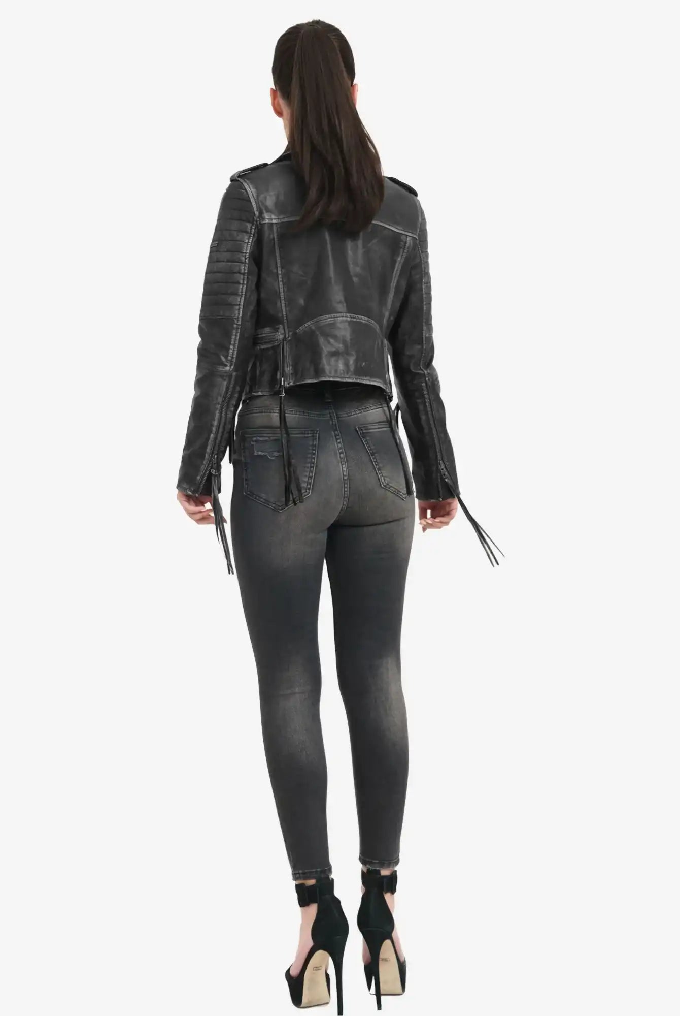Eine Frau mit langen dunklen Haaren in einem Pferdeschwanz steht mit dem Rücken zur Kamera. Sie trägt die trueprodigy Avril Bikerlederjacke Vintage Optik, verwaschene schwarze Skinny Jeans und stylische schwarze hochhackige Schuhe.
