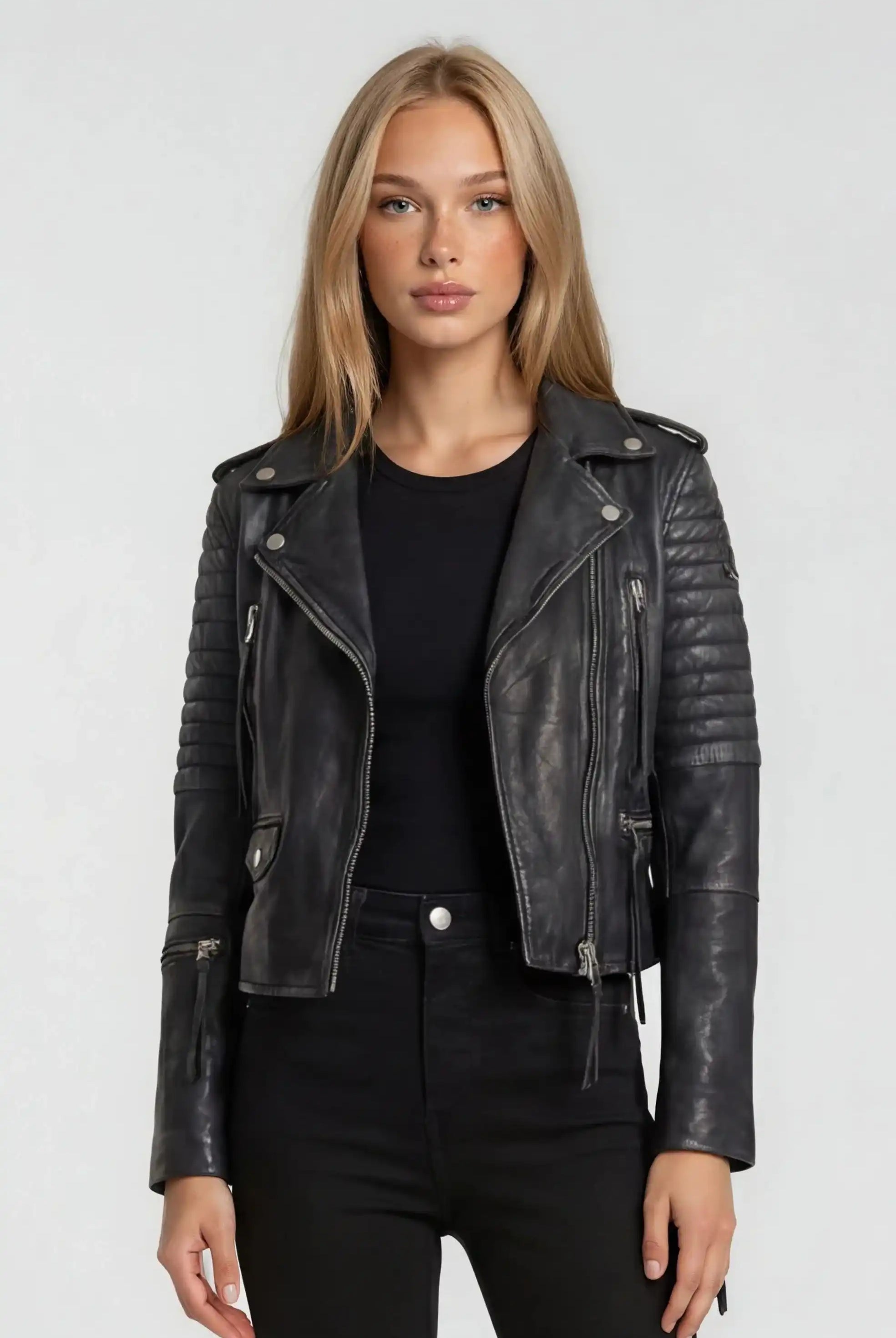 Eine Frau mit glattem blondem Haar trägt die trueprodigy Bikerlederjacke Vintage Optik über einem schwarzen Oberteil und einer Hose, steht vor einem schlichten hellgrauen Hintergrund und schaut direkt in die Kamera.