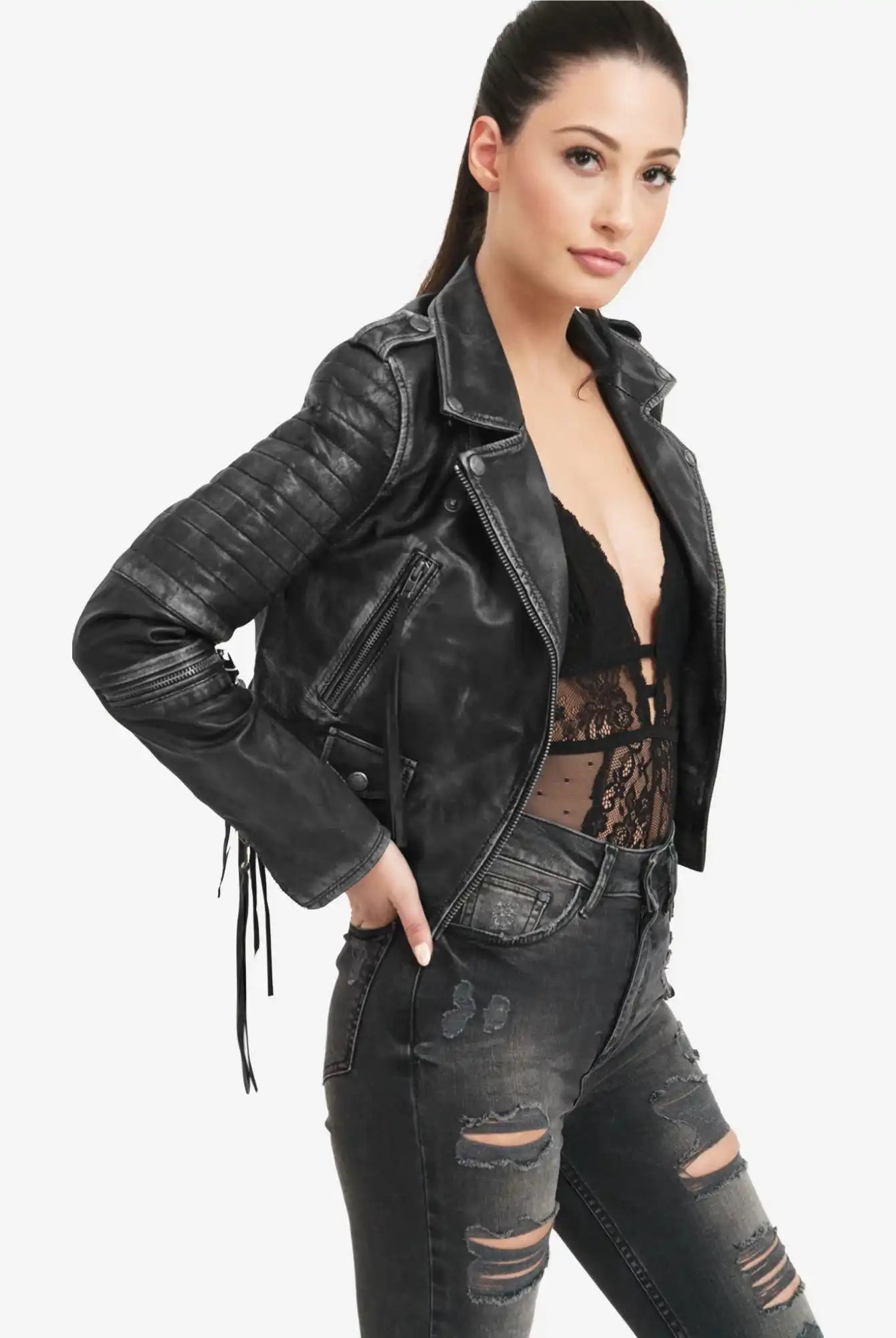 Eine Frau trägt die trueprodigy Avril Bikerlederjacke Vintage Optik mit einem schwarzen Spitzentop und einer schwarzen Jeans im Used-Look und steht mit einer Hand an der Hüfte vor einem schlichten weißen Hintergrund.