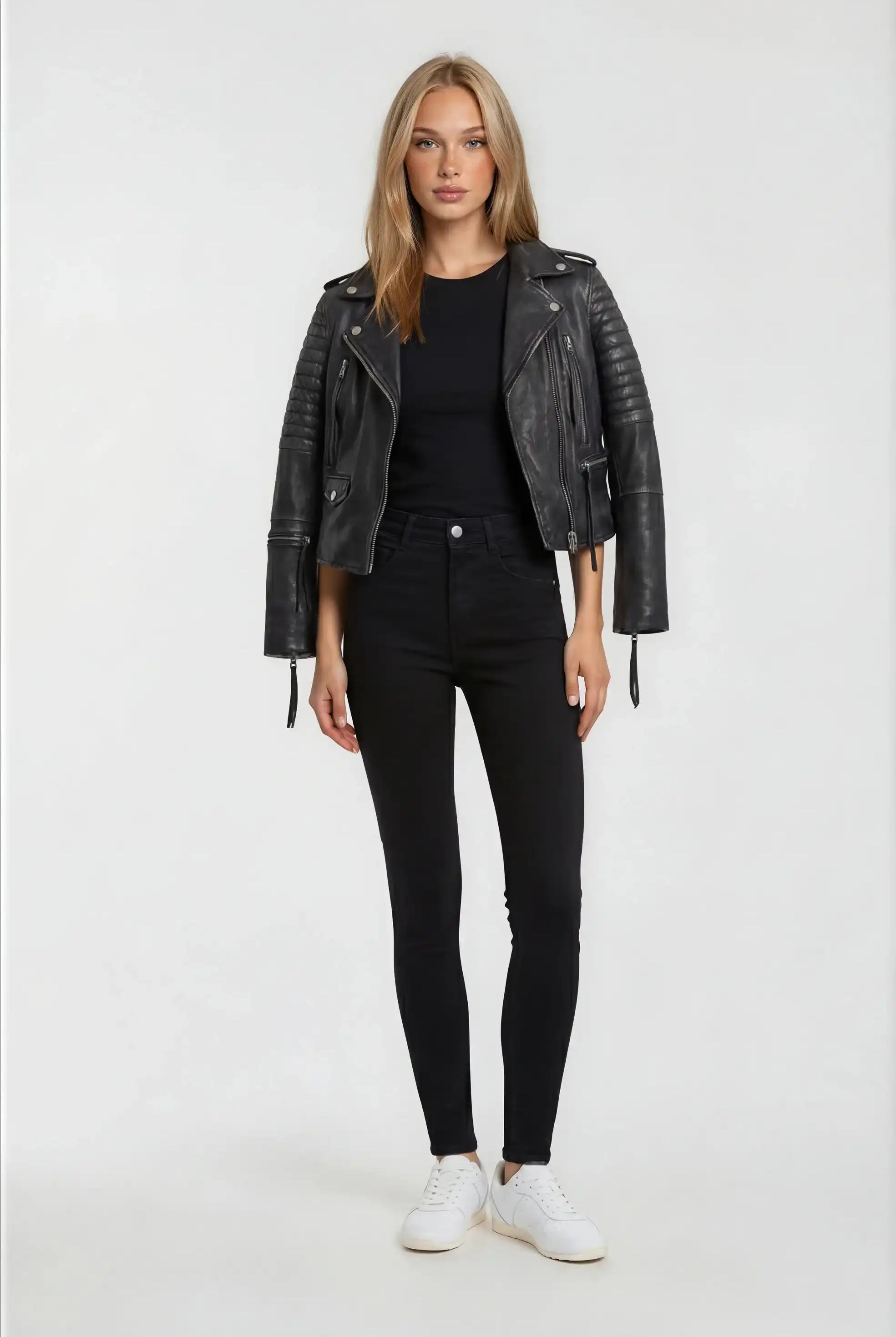 Eine Frau mit glattem blondem Haar und neutralem Gesichtsausdruck blickt nach vorne. Sie trägt die trueprodigy Bikerlederjacke Vintage Optik in Schwarz, ein schwarzes Hemd, High-Waisted Skinny Jeans und weiße Turnschuhe vor einem schlichten hellen Hintergrund.
