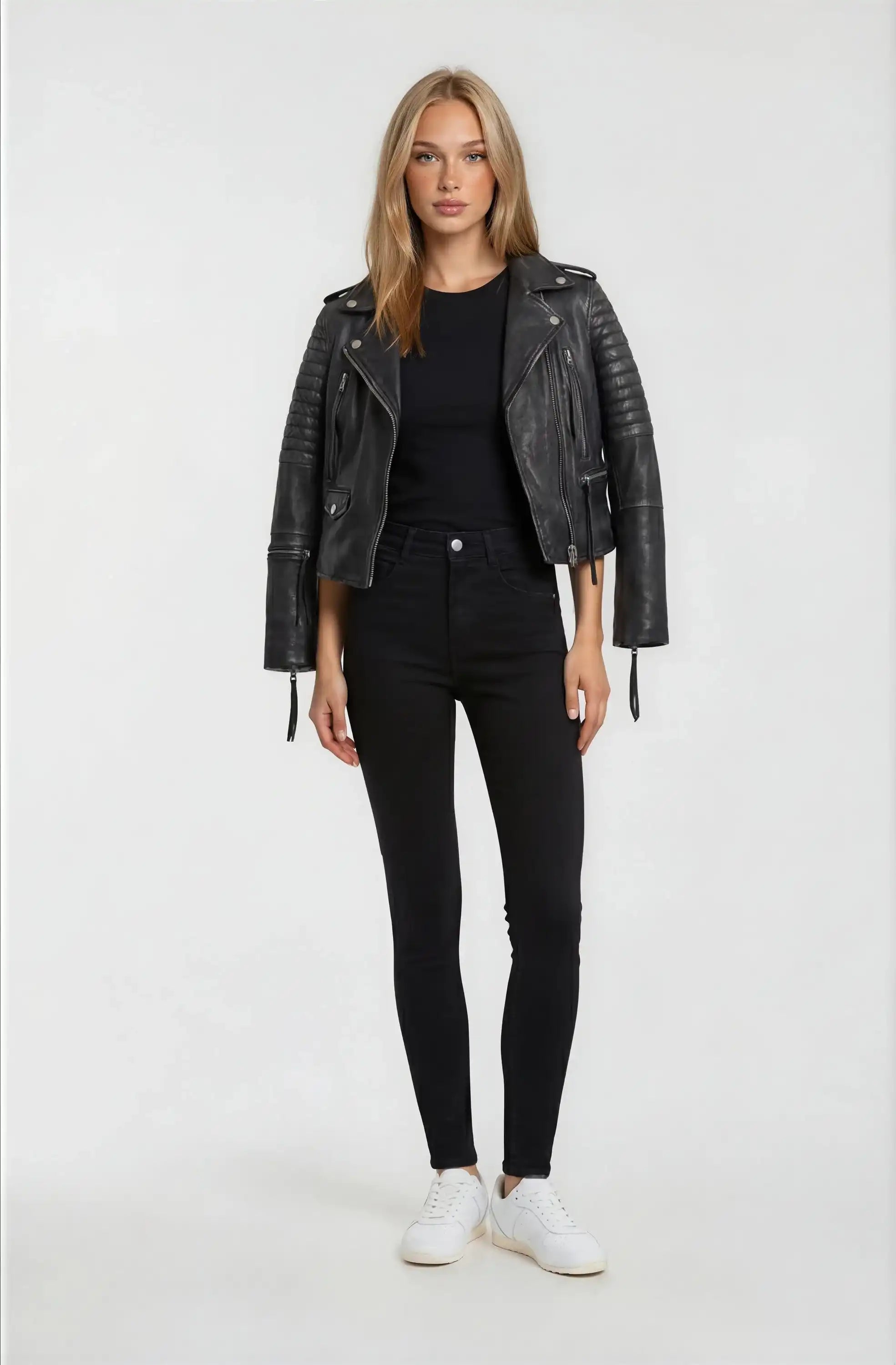 Eine Frau mit glattem blondem Haar und neutralem Gesichtsausdruck blickt nach vorne. Sie trägt die trueprodigy Bikerlederjacke Vintage Optik in Schwarz, ein schwarzes Hemd, High-Waisted Skinny Jeans und weiße Turnschuhe vor einem schlichten hellen Hintergrund.