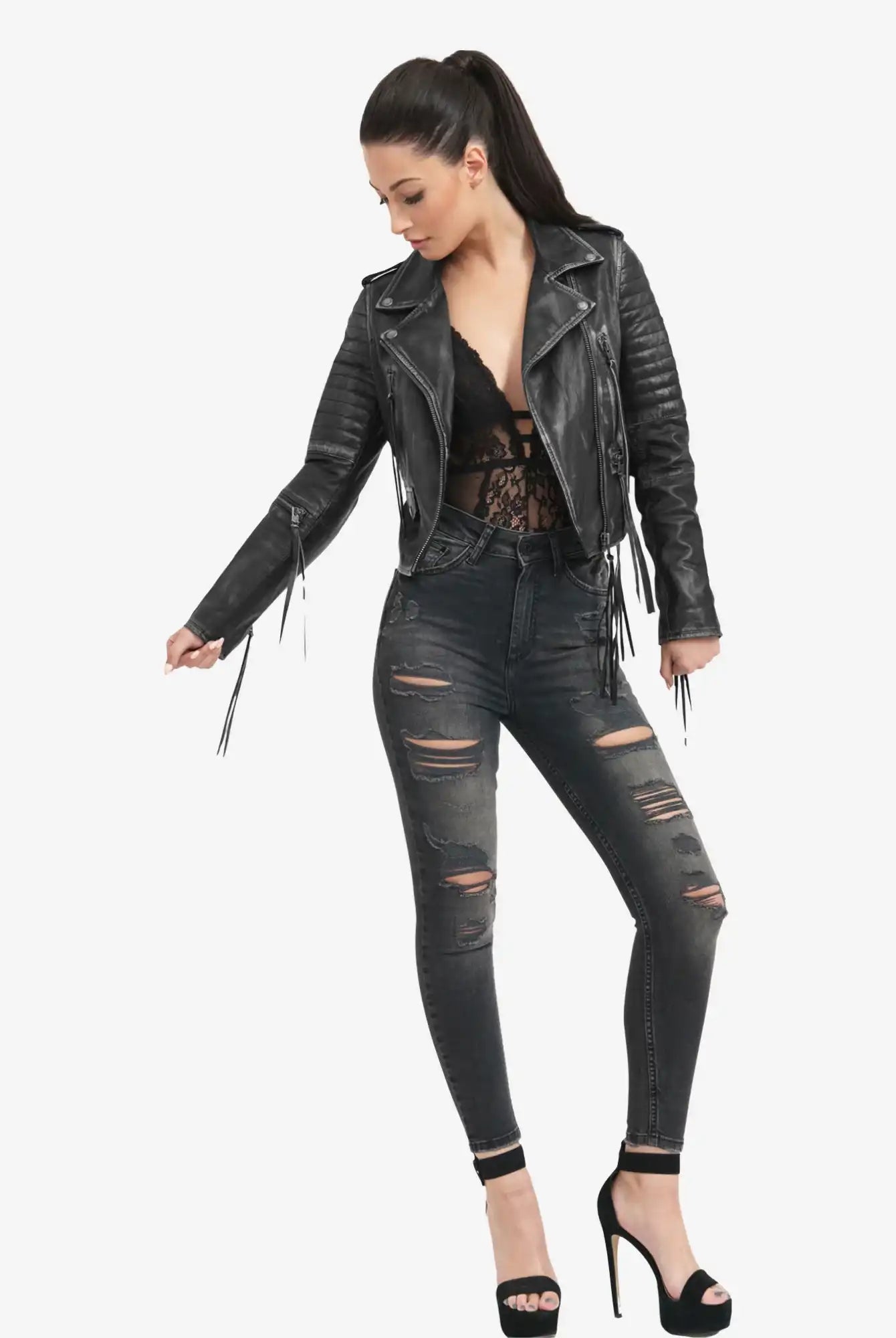 Eine Frau mit langen dunklen Haaren in einem Pferdeschwanz trägt die Avril Bikerlederjacke Vintage Optik von trueprodigy in Schwarz, gepaart mit einem schwarzen Spitzentop, zerrissenen dunklen Skinny Jeans und schwarzen hochhackigen Sandalen, vor einem schlichten weißen Hintergrund.