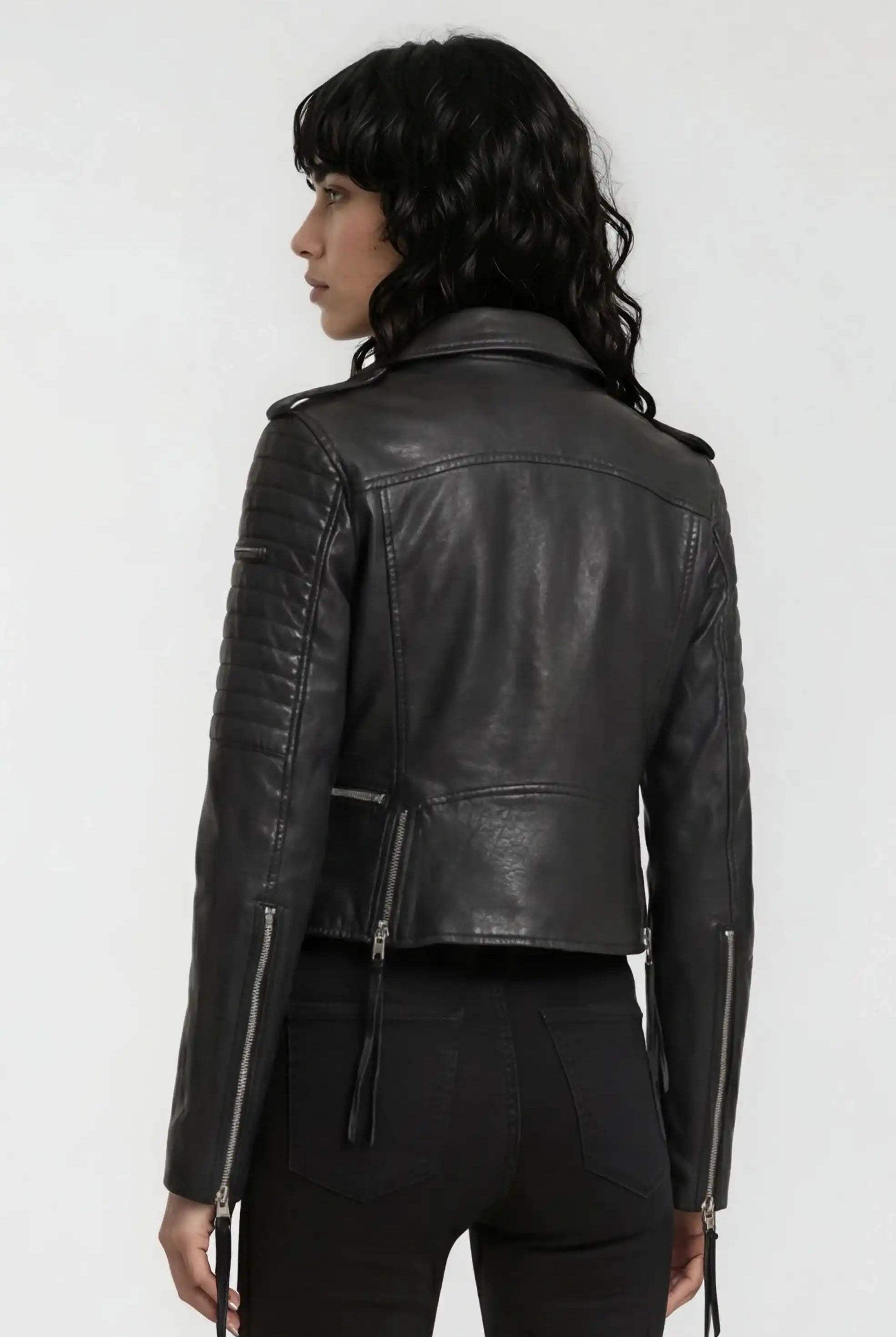 Eine Person mit gewellten dunklen Haaren ist von hinten zu sehen. Sie trägt die trueprodigy Biker Lederjacke und eine schwarze Hose und steht vor einem schlichten hellen Hintergrund.