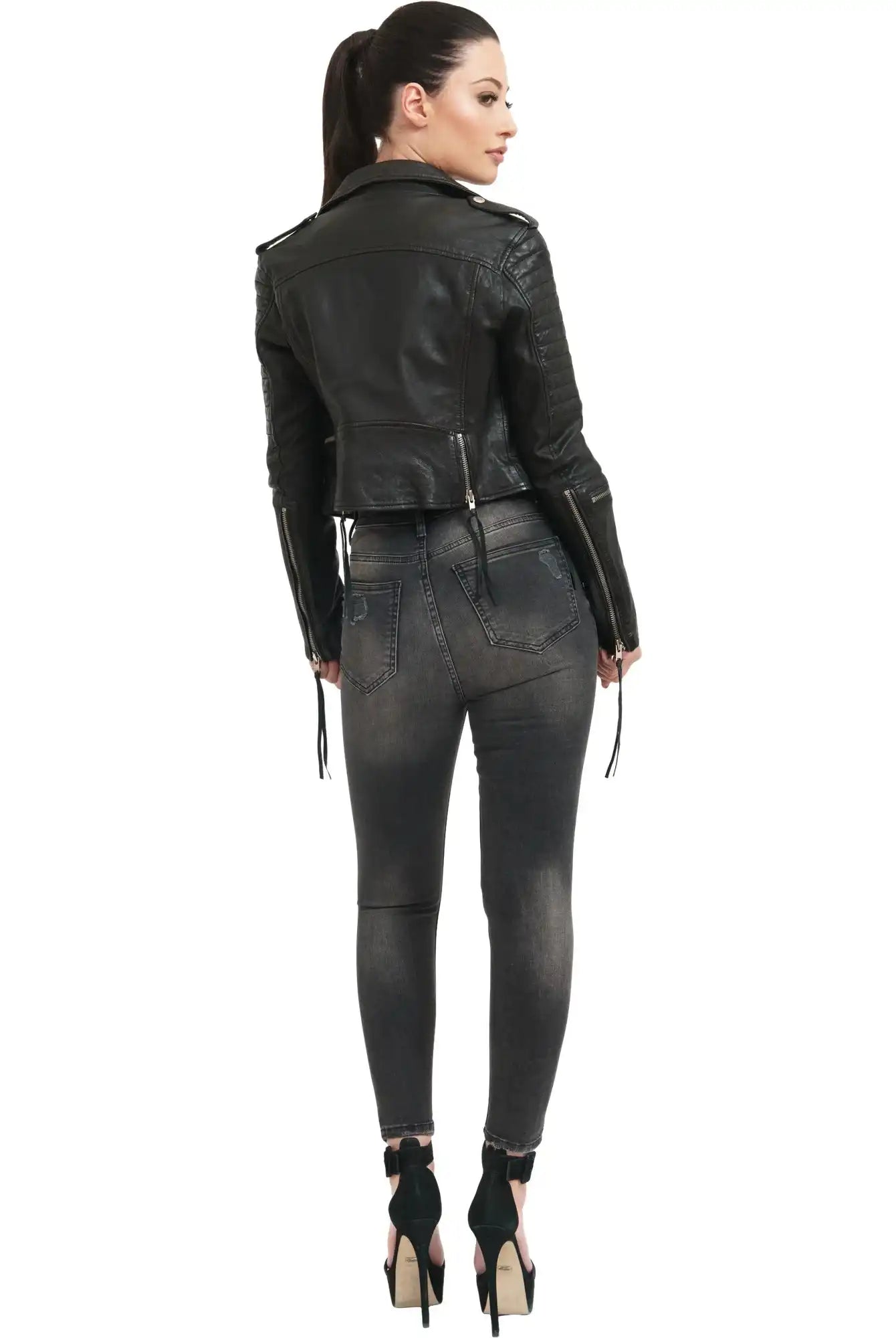 Eine Frau mit Pferdeschwanz steht mit dem Gesicht zur Wand und trägt die trueprodigy Jade Biker Lederjacke, verwaschene schwarze Skinny Jeans und schwarze hochhackige Schuhe vor einem schlichten weißen Hintergrund.