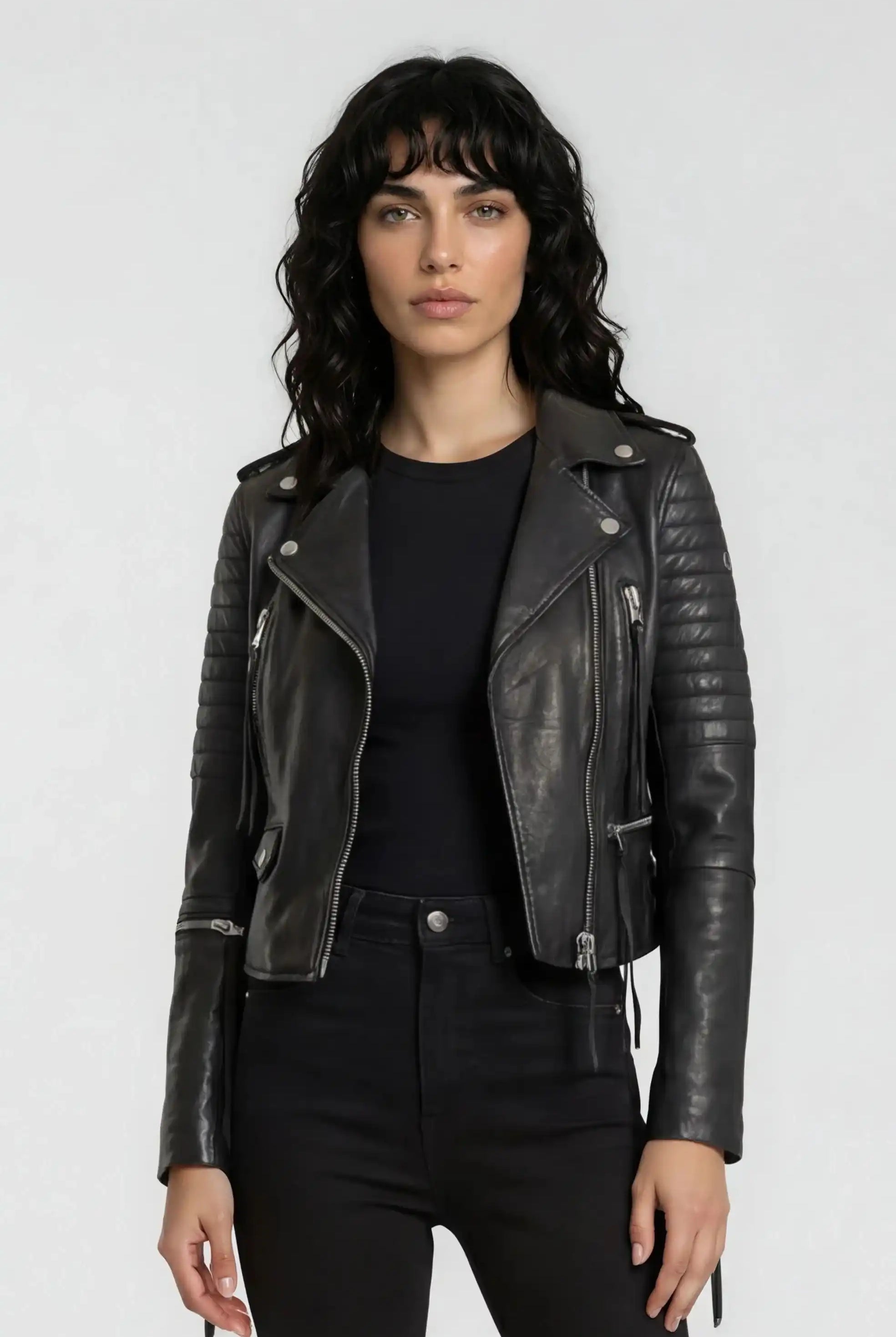 Eine Frau mit gewelltem dunklem Haar trägt eine trueprodigy Biker Lederjacke über einem schwarzen Hemd und einer Hose. Sie steht vor einem schlichten hellgrauen Hintergrund und schaut mit neutralem Blick in die Kamera.