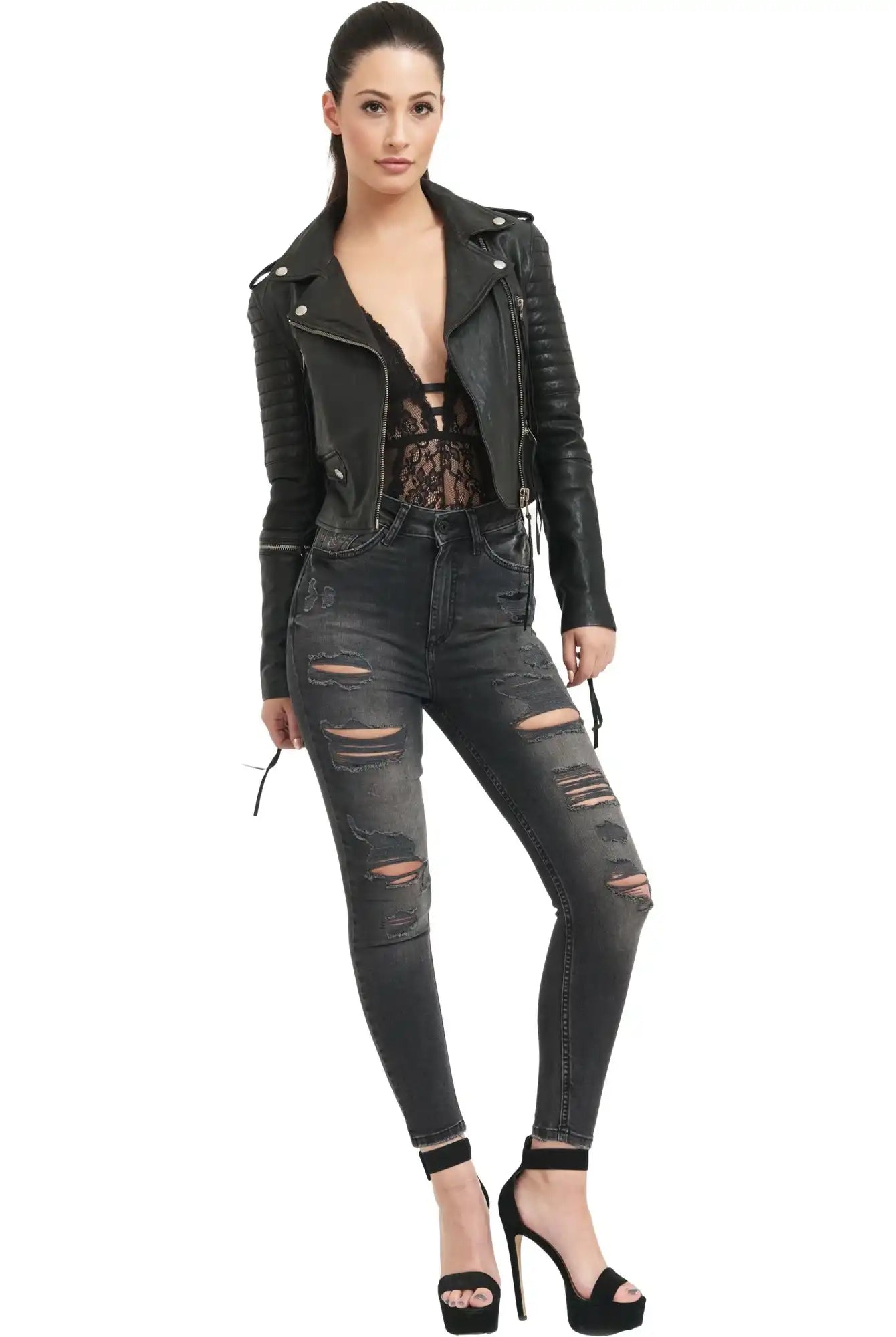 Eine Frau posiert vor einem weißen Hintergrund und trägt die trueprodigy Jade Biker Lederjacke mit einem schwarzen Spitzentop, zerrissenen Jeans und hochhackigen Sandalen. Ihr dunkles Haar ist zurückgesteckt und eine Hand ruht lässig in ihrer Tasche.