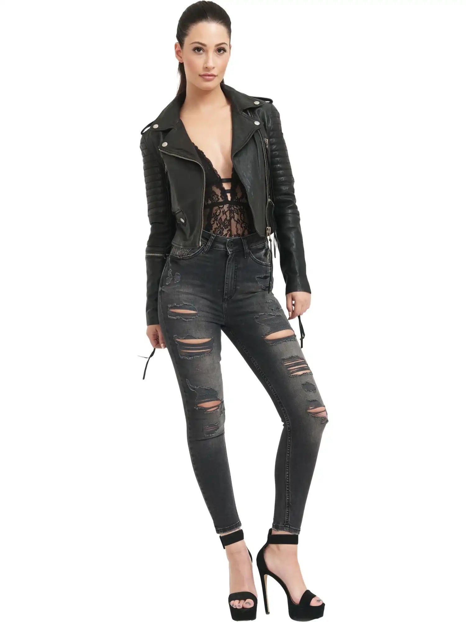 Eine Frau posiert vor einem weißen Hintergrund und trägt die trueprodigy Jade Biker Lederjacke mit einem schwarzen Spitzentop, zerrissenen Jeans und hochhackigen Sandalen. Ihr dunkles Haar ist zurückgesteckt und eine Hand ruht lässig in ihrer Tasche.