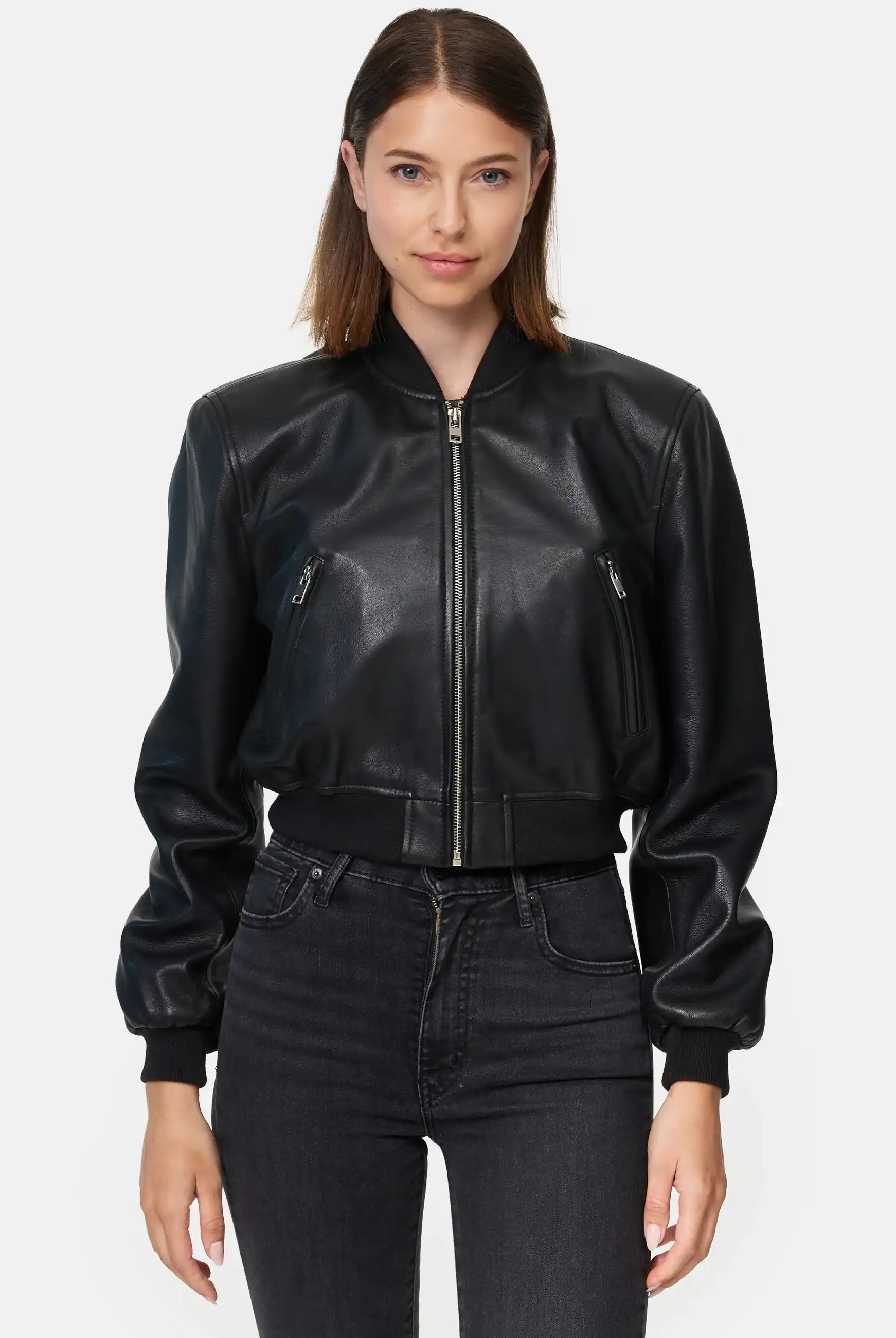 Eine Frau steht mit dem Blick nach vorne und trägt die trueprodigy Paola Kurze Oversized Lederjacke, eine schwarze Lederjacke mit Reißverschluss, gepaart mit einer schwarzen High-Waisted-Jeans vor einem schlichten hellen Hintergrund.