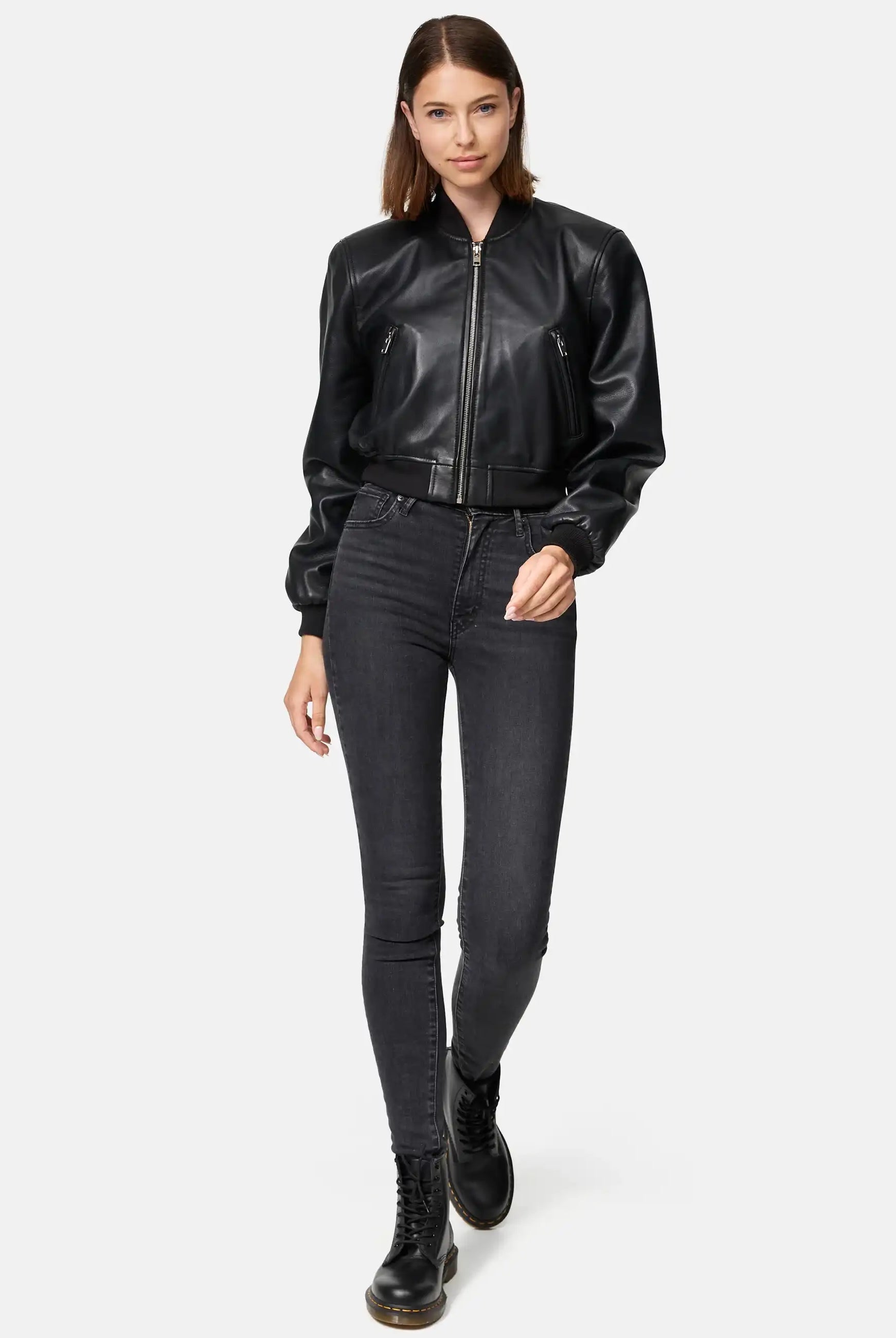 Eine Frau mit glattem braunem Haar trägt eine Trueprodigy Paola Kurze Oversized Lederjacke, dunkle High-Waisted Skinny Jeans und schwarze Kampfstiefel. Sie steht vor einem schlichten hellen Hintergrund und schaut mit neutralem Blick in die Kamera.