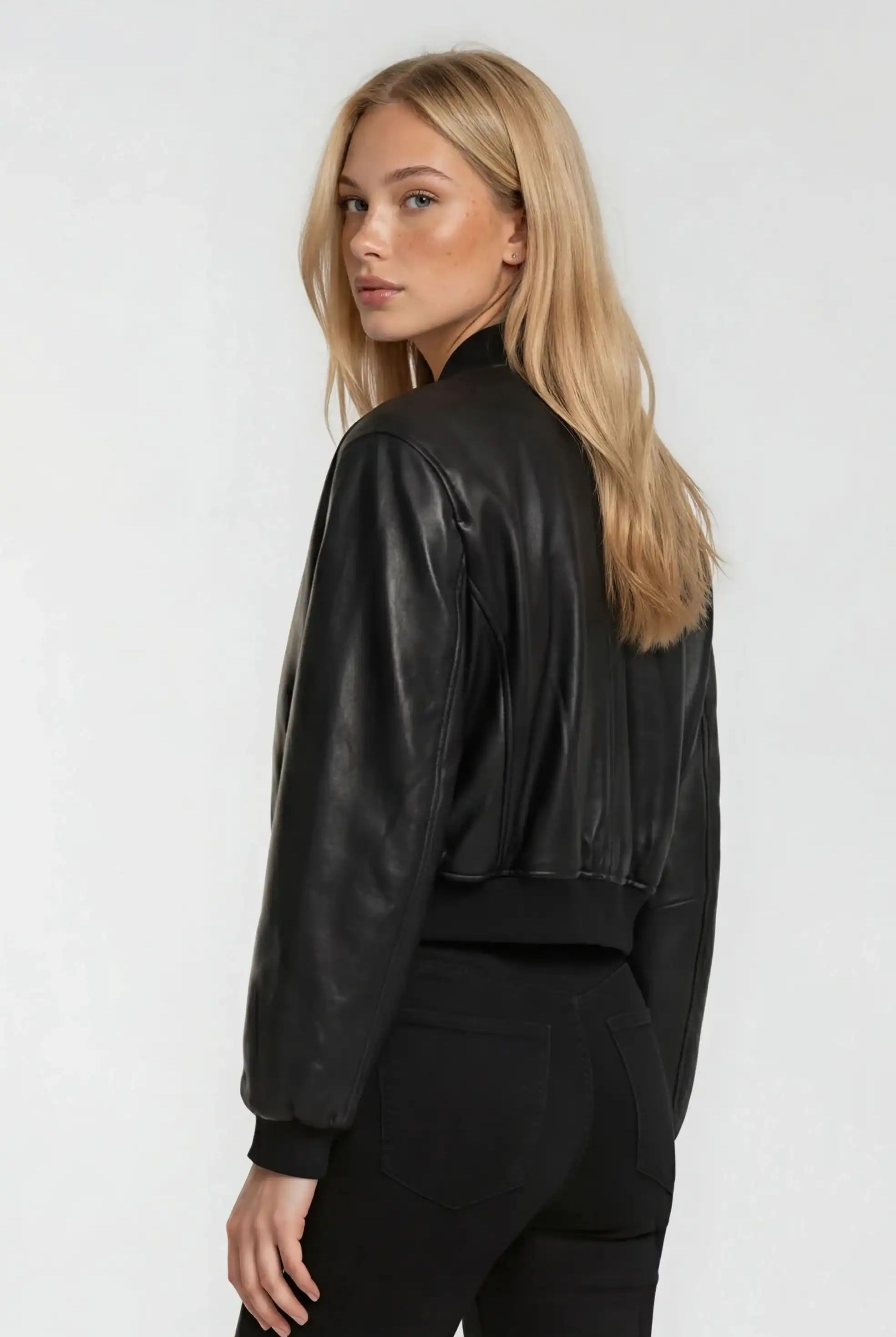 Eine Frau mit langen blonden Haaren steht vor einem hellen Hintergrund und schaut über ihre Schulter. Sie trägt eine schwarze Hose und die Kurze Oversized Lederjacke von trueprodigy.