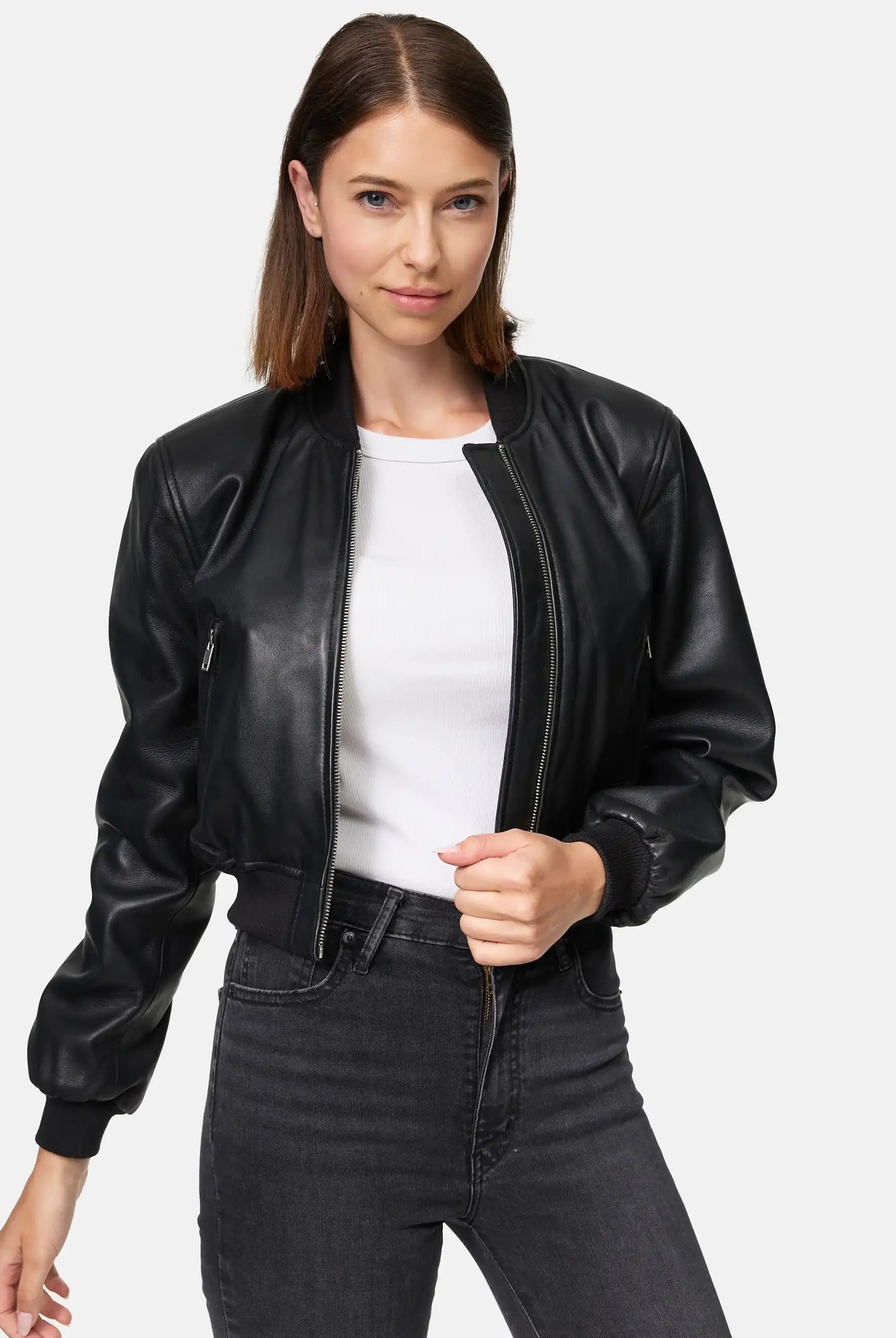 Eine Frau trägt die Paola Kurze Oversized Lederjacke von trueprodigy über einem weißen Shirt und dunklen Jeans. Sie steht vor einem schlichten Hintergrund, hat glattes braunes Haar und hält den Jackenreißverschluss mit einer Hand.