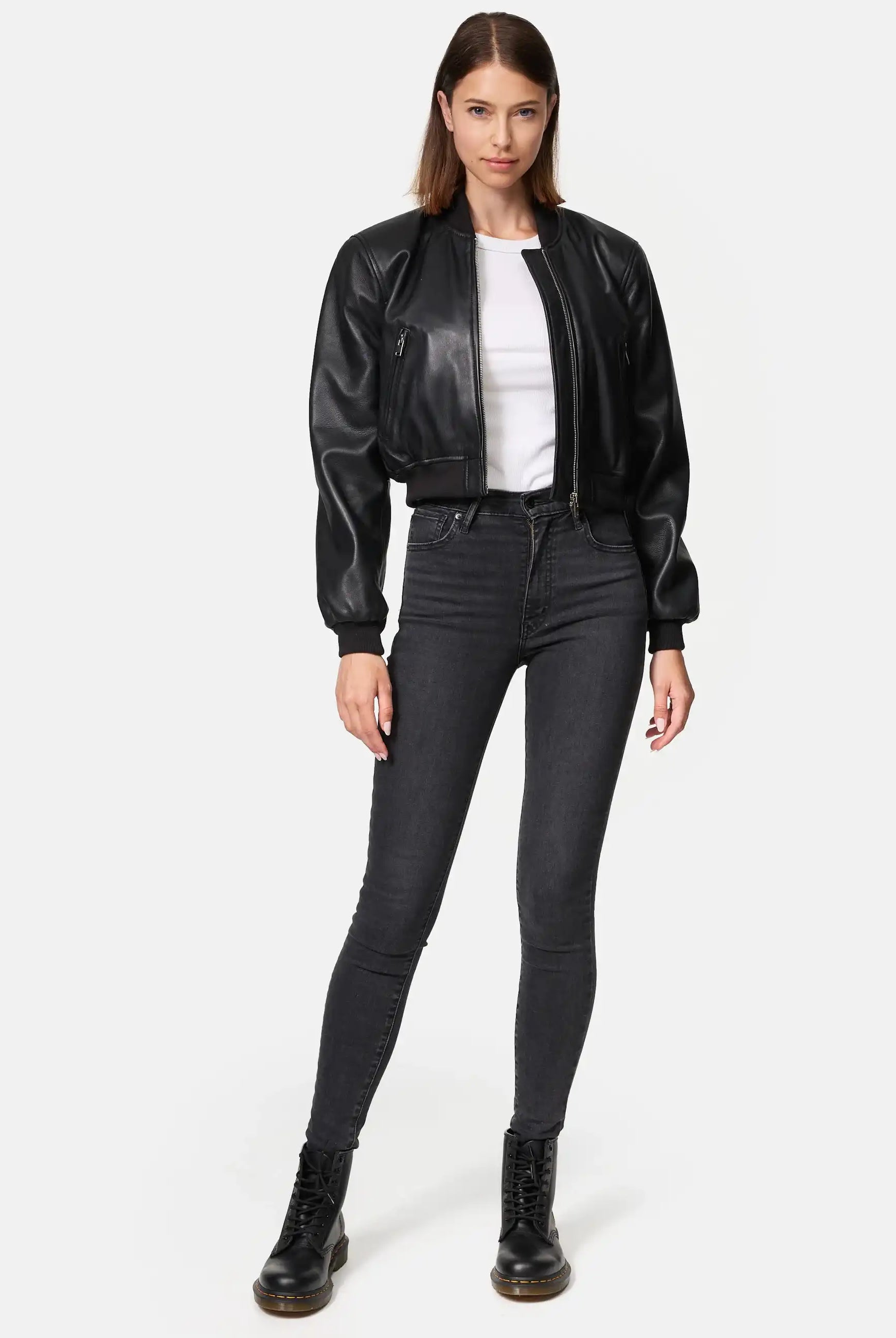 Eine Frau steht mit dem Gesicht nach vorne in einer Paola Kurze Oversized Lederjacke, einem weißen T-Shirt, einer schwarzen Jeans mit hoher Taille und schwarzen Schnürstiefeln vor einem schlichten weißen Hintergrund.